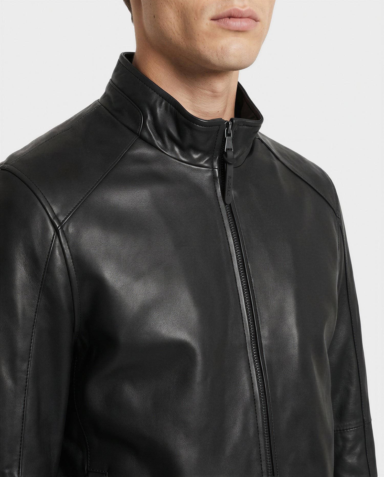 A person wearing the Rudsak GARY - M126061 Black leather jacket, featuring a high collar and front zipper, shown from shoulders to chin against a plain background.