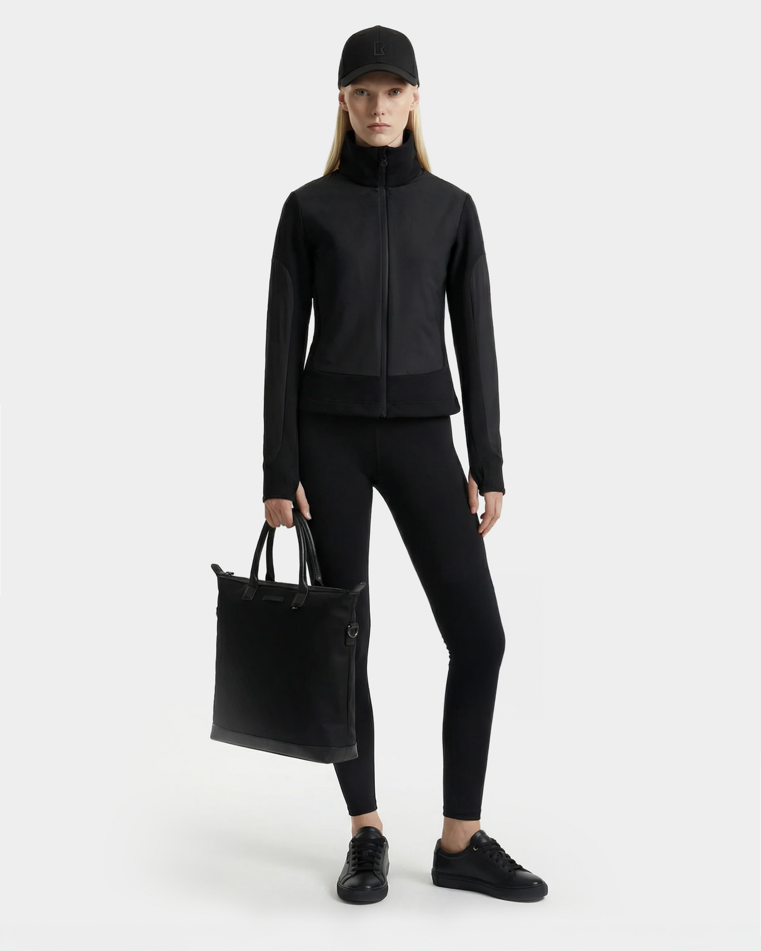 A woman stands against a plain background in an all-black Rudsak outfit, holding the GRANADA - 8126023 Black tote bag in her left hand and looking directly at the camera.