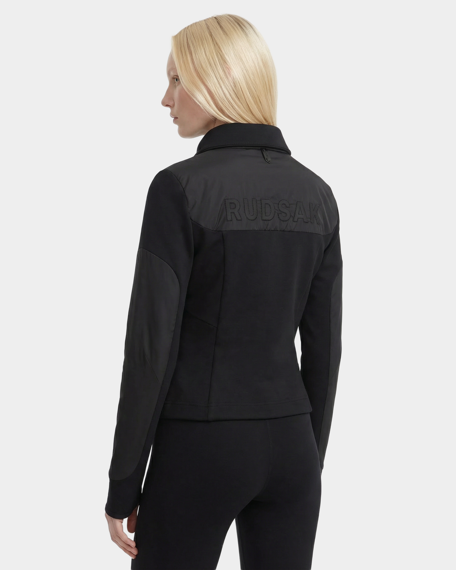 A person with long blonde hair faces away from the camera, wearing the Rudsak GRANADA - 8126023 Black sporty-chic sweater jacket with "RUDSAK" embroidered on the back, set against a plain white background.