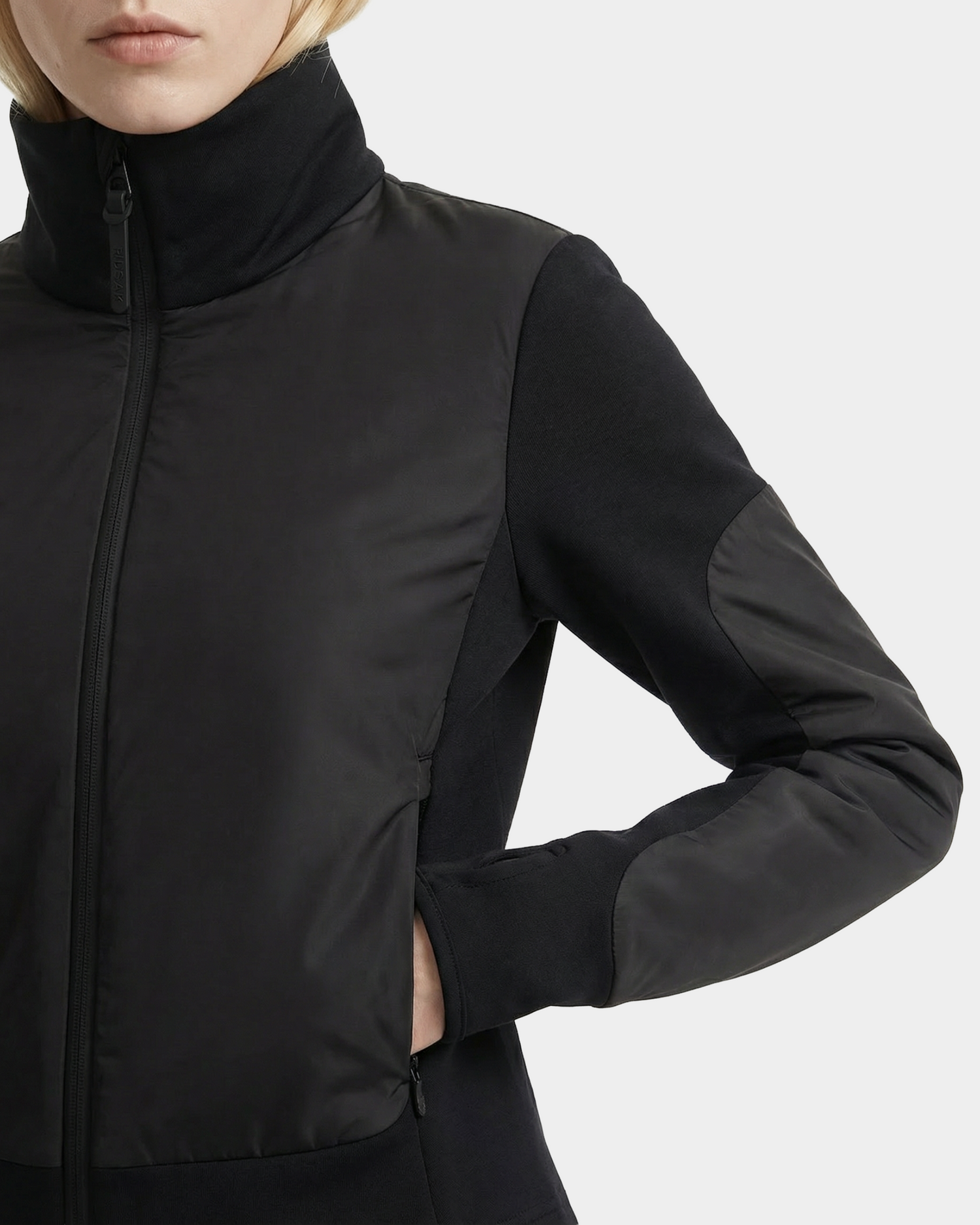A person models the Rudsak GRANADA - 8126023 Black zip-up sweater jacket, featuring a high collar, textured sleeves and chest, with one hand in a side pocket. Cropped at the lips and waist to showcase this sporty-chic layer.