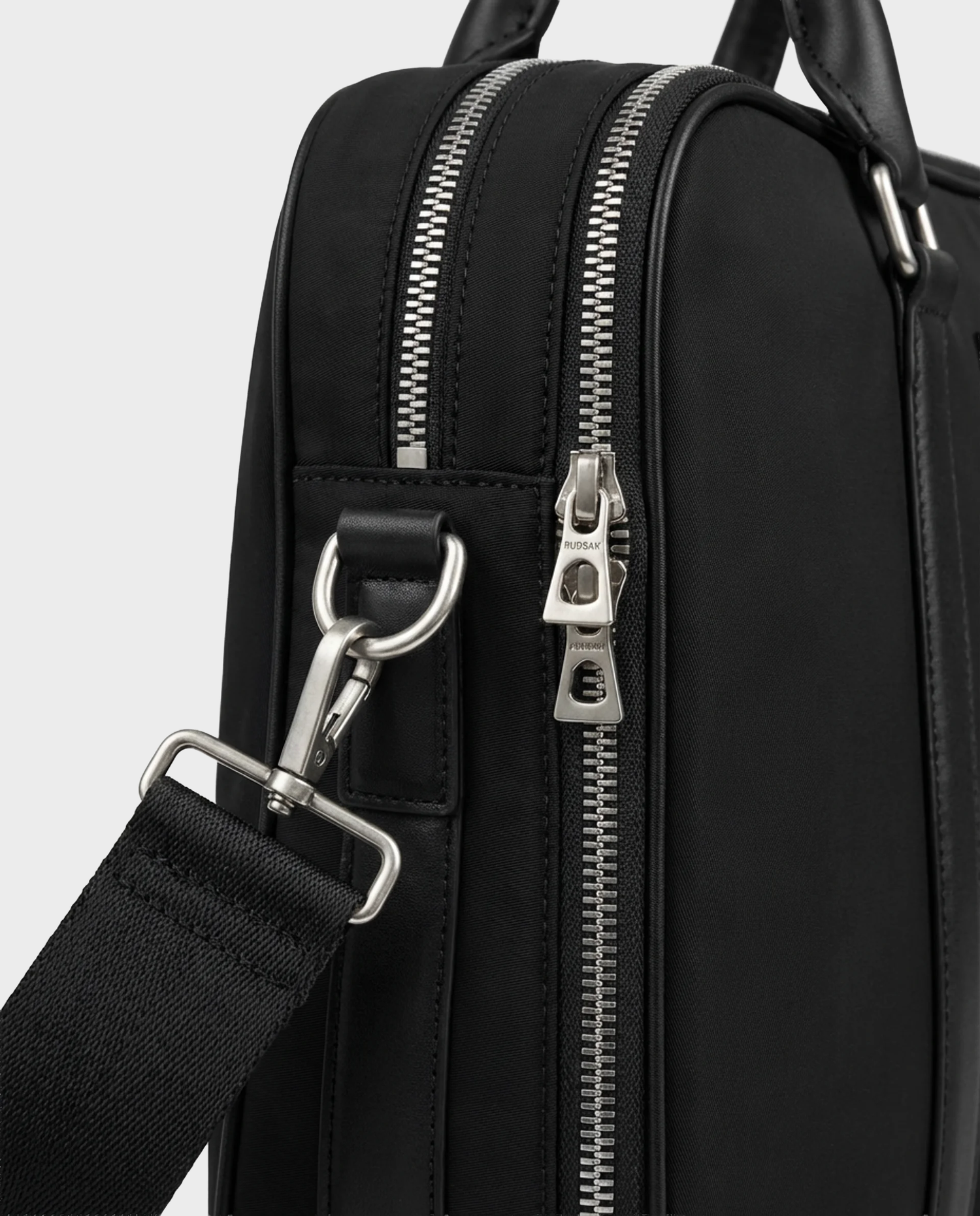 Close-up of the Rudsak HENRY N - 9326056 Black bag, featuring silver zippers and hardware with a detachable black strap clipped to a metal ring, highlighting the bag’s texture and stitching details.
