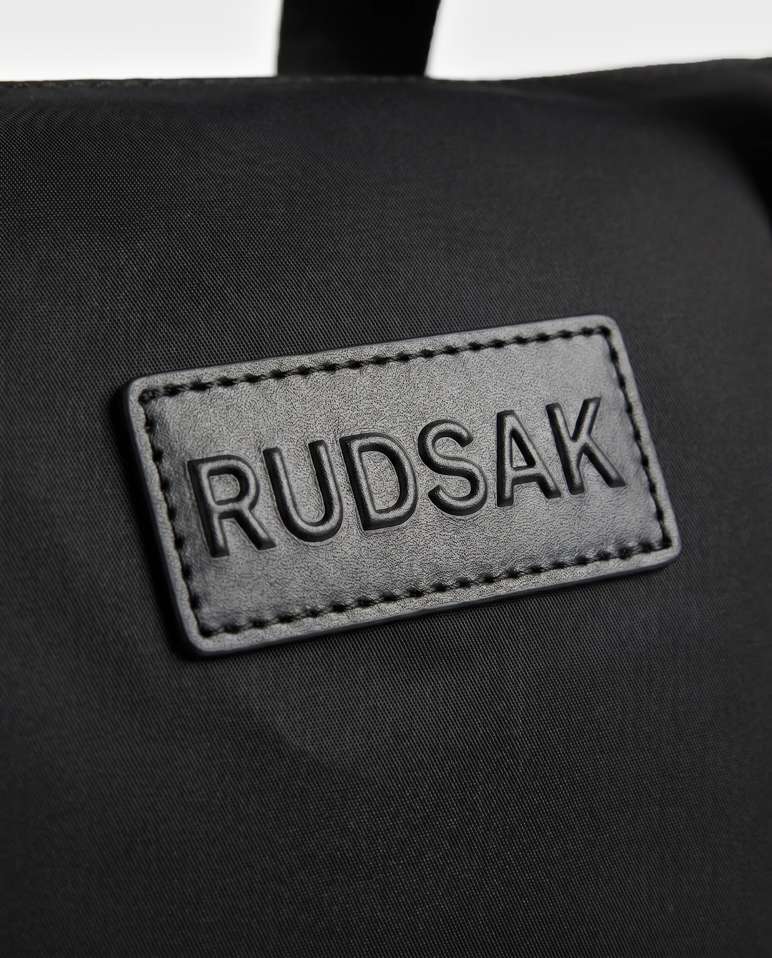 Detailed shot of a black fabric patch stitched onto the HENRY N - 9326056 Black bag, featuring the Rudsak brand name embossed in capital letters.