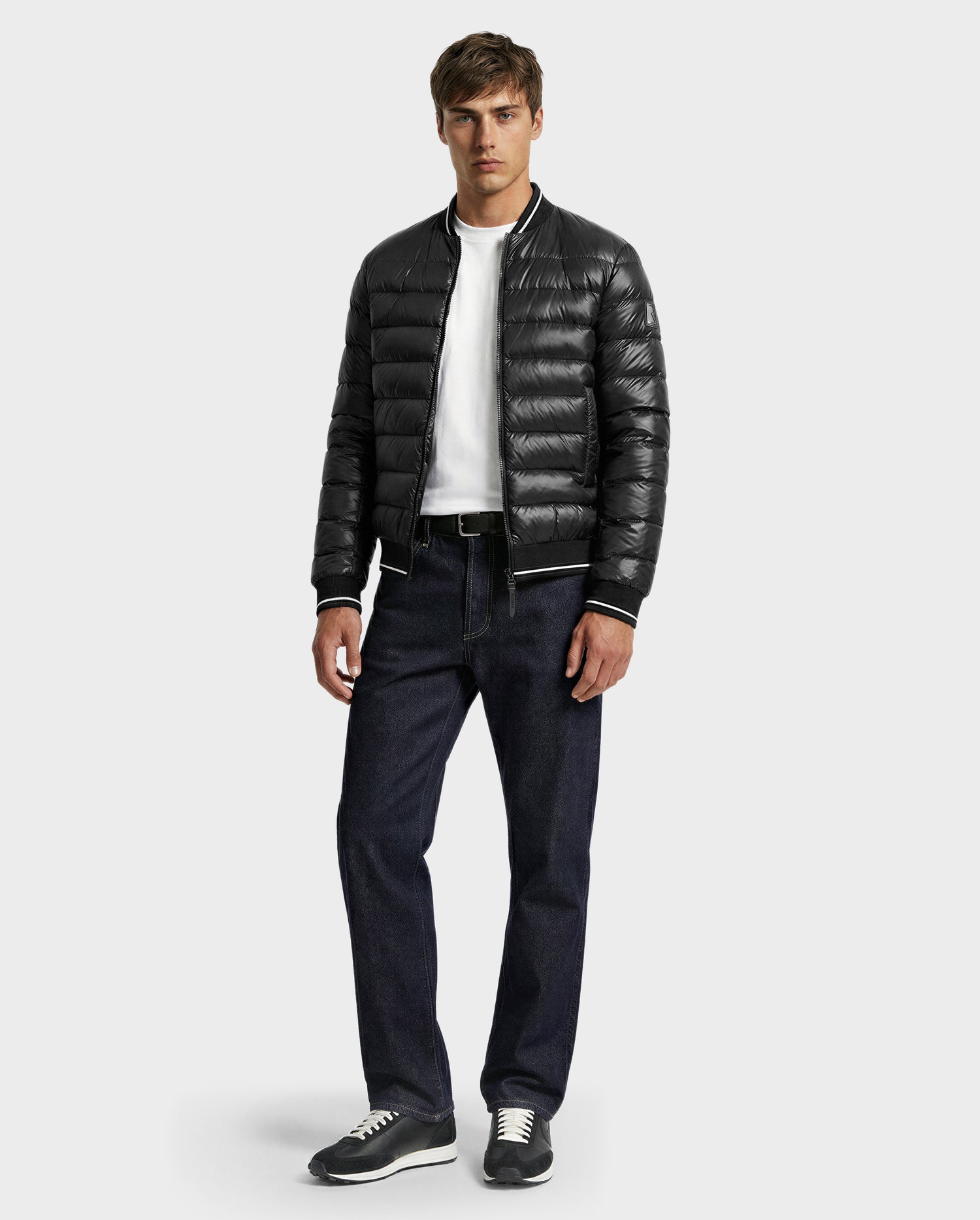 A man stands facing forward, wearing the Rudsak JEREMY - 6125025 Black down bomber jacket over a white shirt, dark blue jeans, and gray sneakers—an ideal minimalist style against a plain light background.