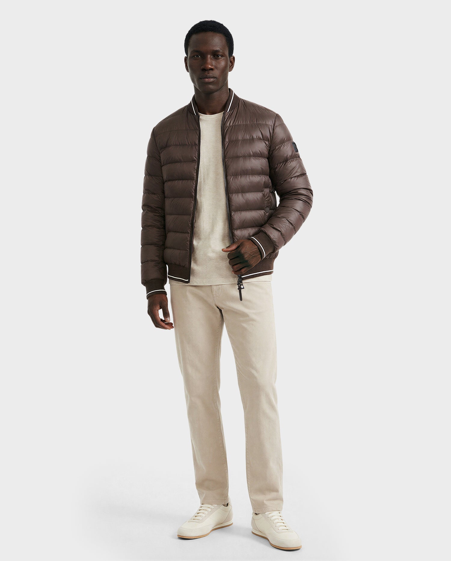 A man wears the Rudsak JEREMY - 6125025 Espresso men’s down bomber jacket with premium insulation over a beige sweater, beige pants, and white sneakers—ideal for minimalist wardrobes against a plain light background.