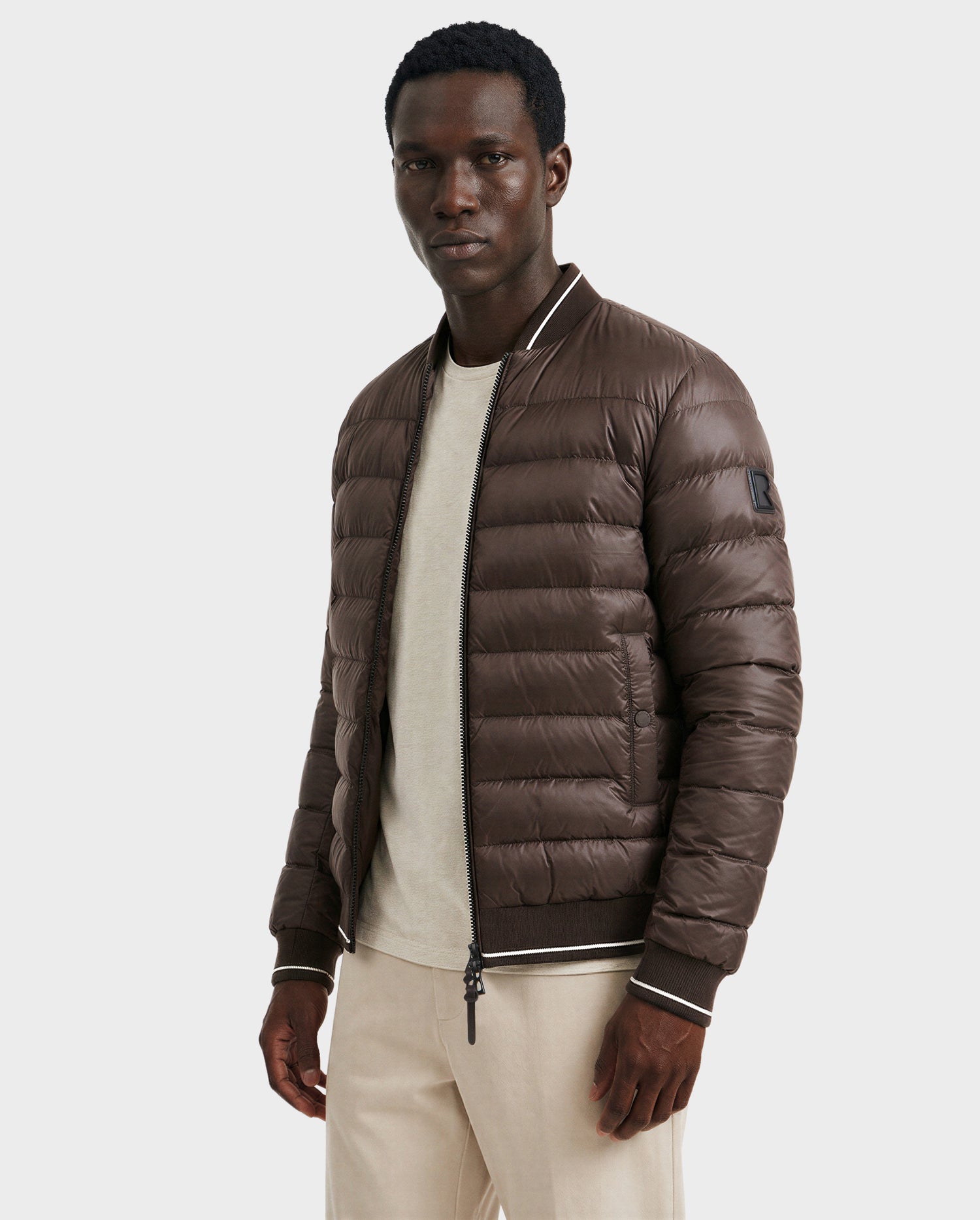 A man wears the Rudsak JEREMY - 6125025 Espresso men's down bomber jacket with premium insulation over a light beige shirt and cream pants, standing against a plain light gray background—ideal for minimalist wardrobes.