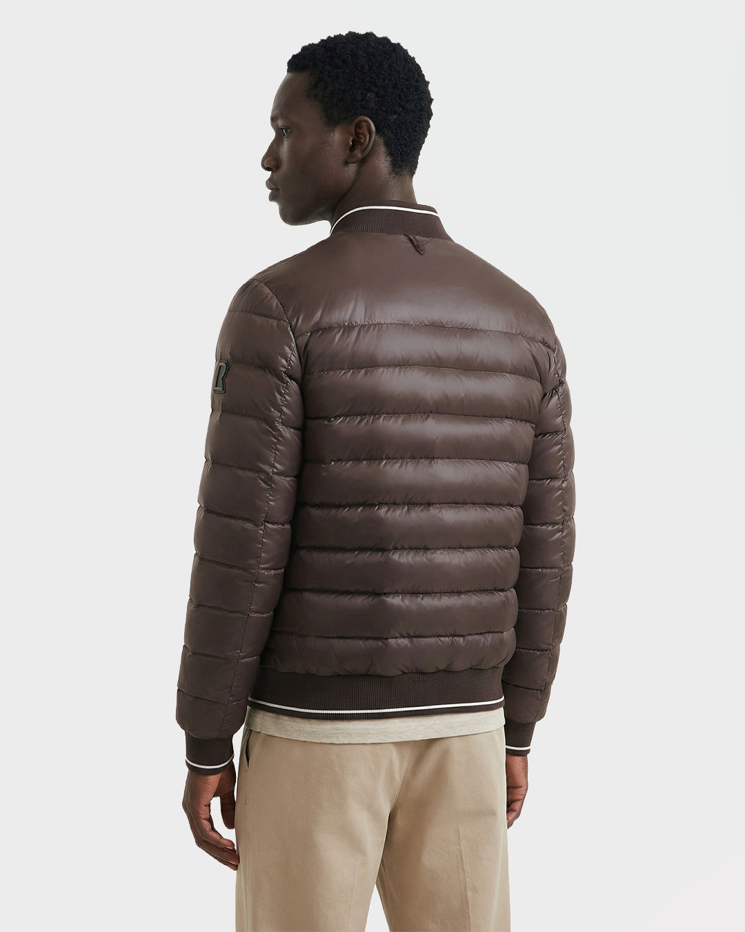 A man stands facing slightly away, wearing the Rudsak JEREMY - 6125025 Espresso men’s down bomber jacket with light trim on the collar and cuffs, paired with beige pants against a plain white background—ideal for minimalist wardrobes.