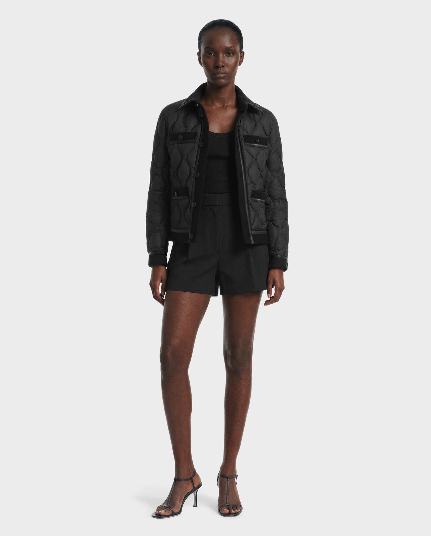 A woman stands facing forward wearing the Rudsak JOLIE - 8125036 Black jacket, black top, black shorts, and black heeled sandals against a plain light gray background.