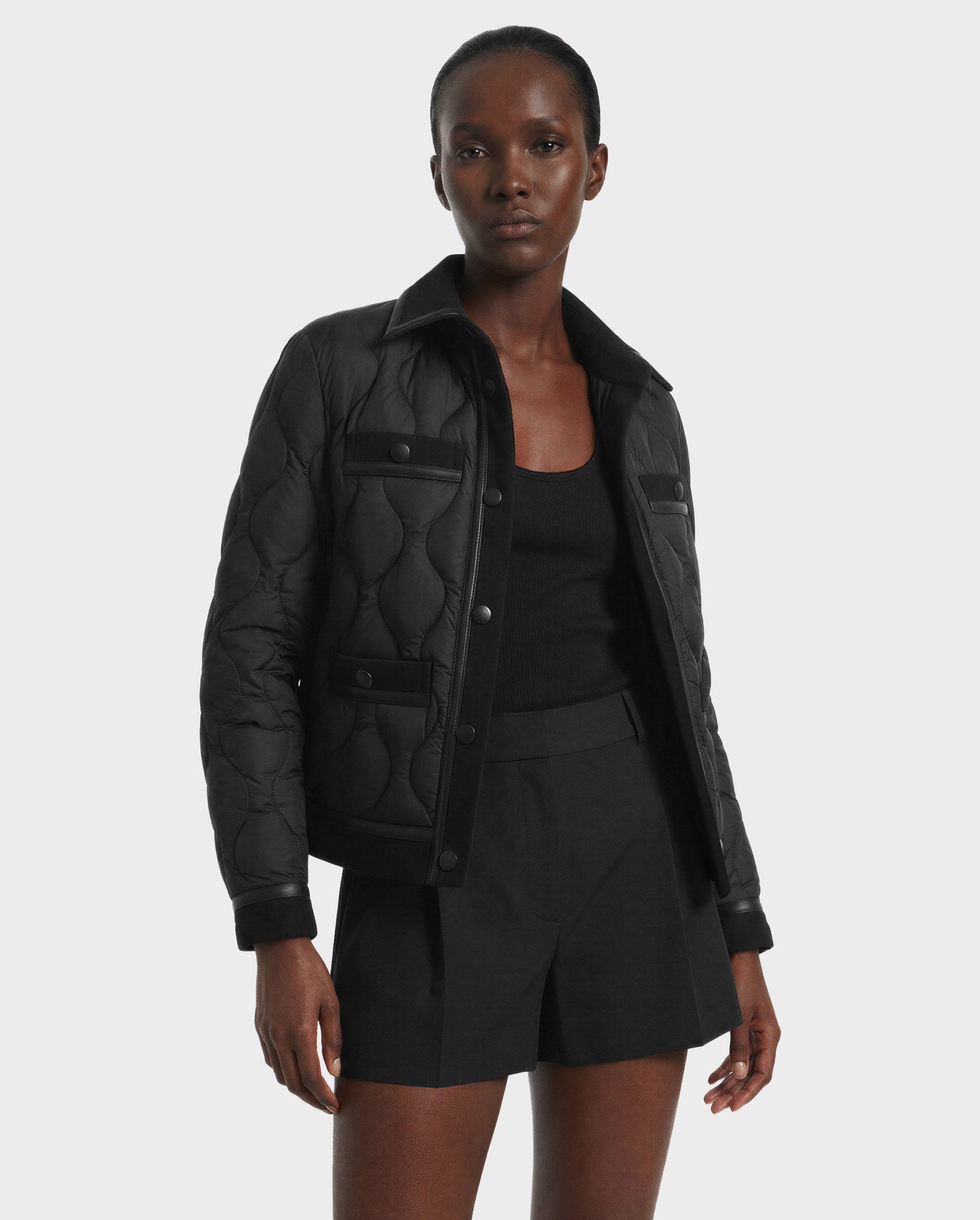 A person wearing the Rudsak JOLIE - 8125036 Black women’s quilted down jacket over a black top and shorts stands against a plain light gray background, looking at the camera.