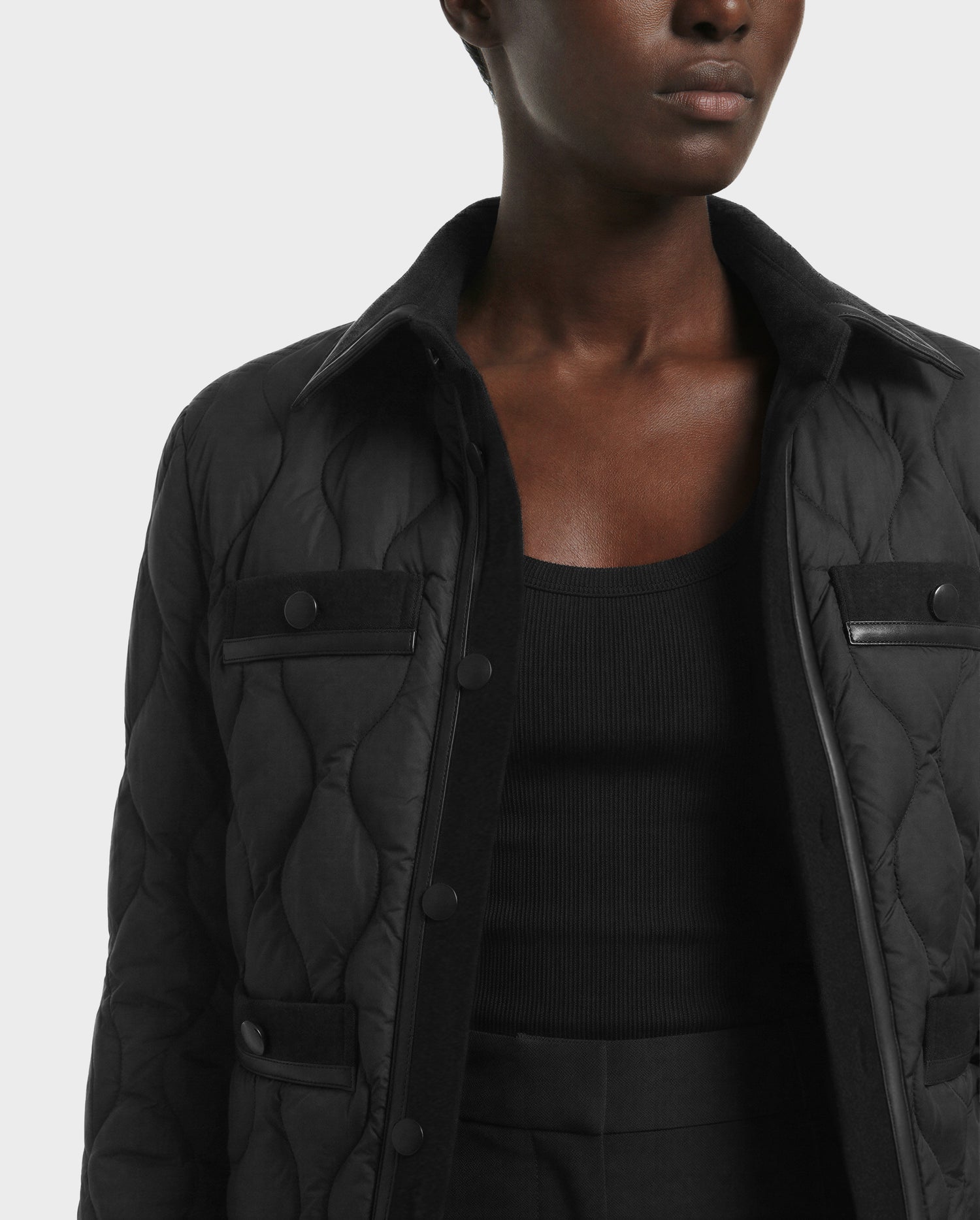 A person wears the Rudsak JOLIE - 8125036 Black women’s quilted down jacket over a black ribbed top, shown from shoulders to waist against a plain light background. The jacket features buttons and flap pockets.