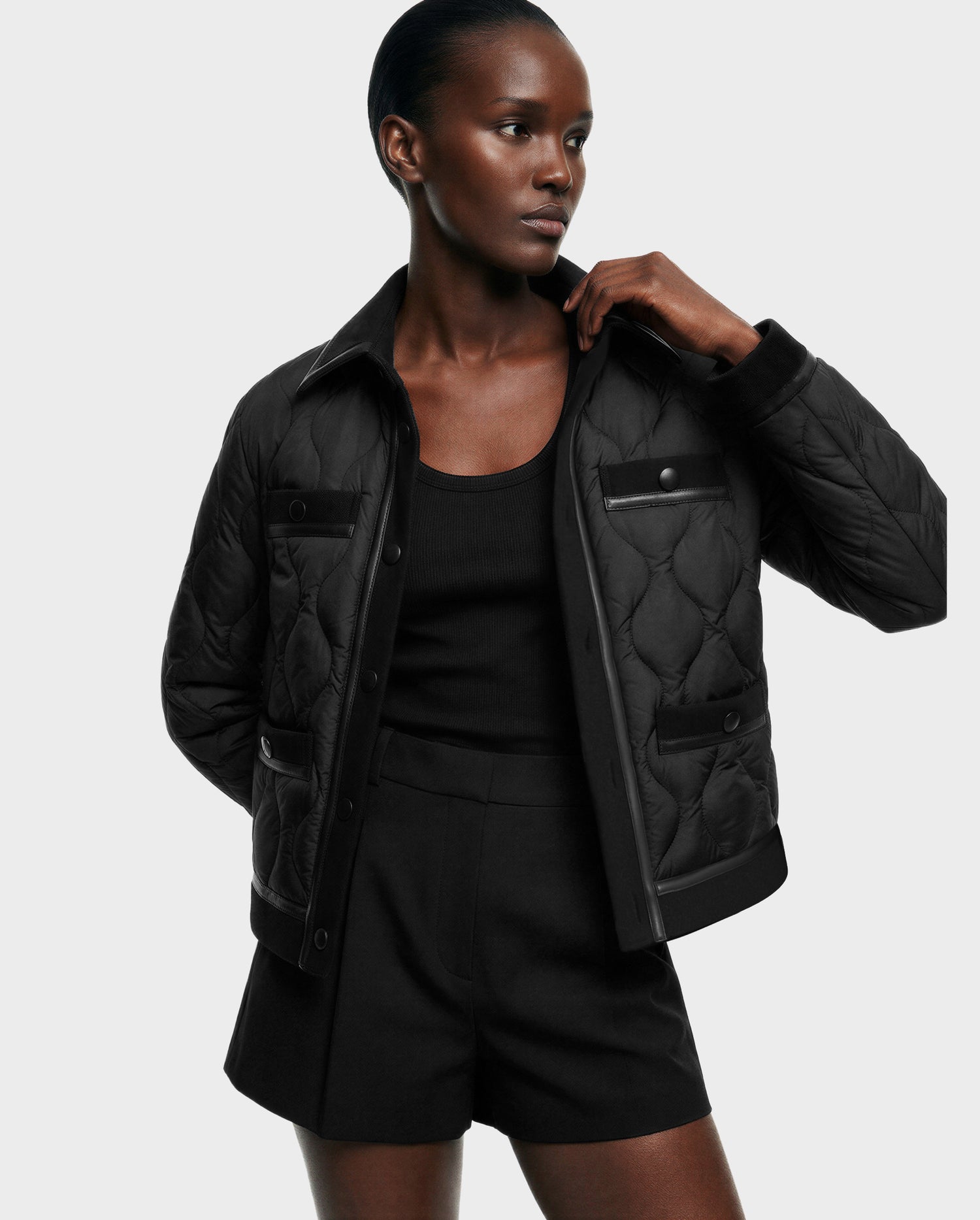 A person models the Rudsak JOLIE - 8125036 Black women’s quilted down jacket over a black tank top and high-waisted shorts, standing against a plain light background with one hand on their collar.