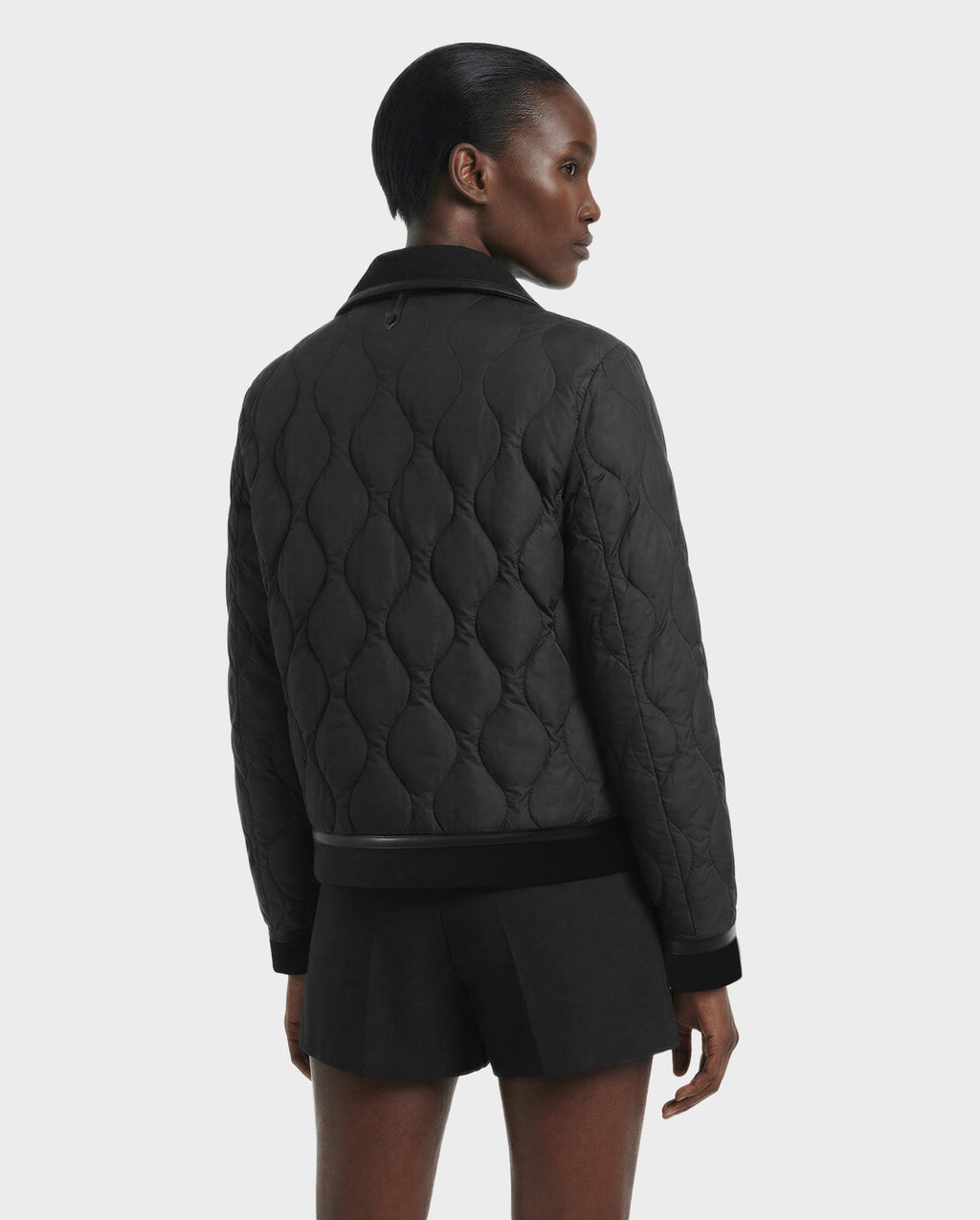 A person with short, slicked-back hair faces away from the camera, wearing the Rudsak JOLIE - 8125036 Black women's quilted down jacket and black shorts, against a plain light gray background.