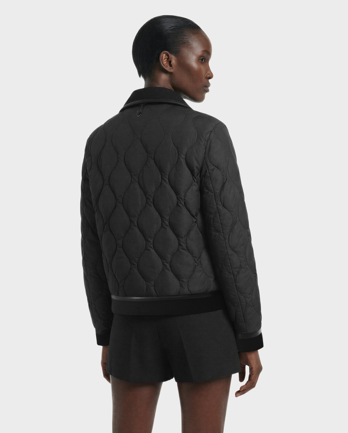 A person with short, slicked-back hair faces away from the camera, wearing the Rudsak JOLIE - 8125036 Black women's quilted down jacket and black shorts, against a plain light gray background.