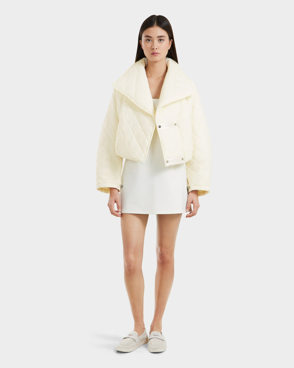 A woman faces forward against a light gray background, wearing the Rudsak KALEE - 8124005 Iceland women’s quilted light jacket with wide lapels over a white mini dress and paired with light-colored loafers.
