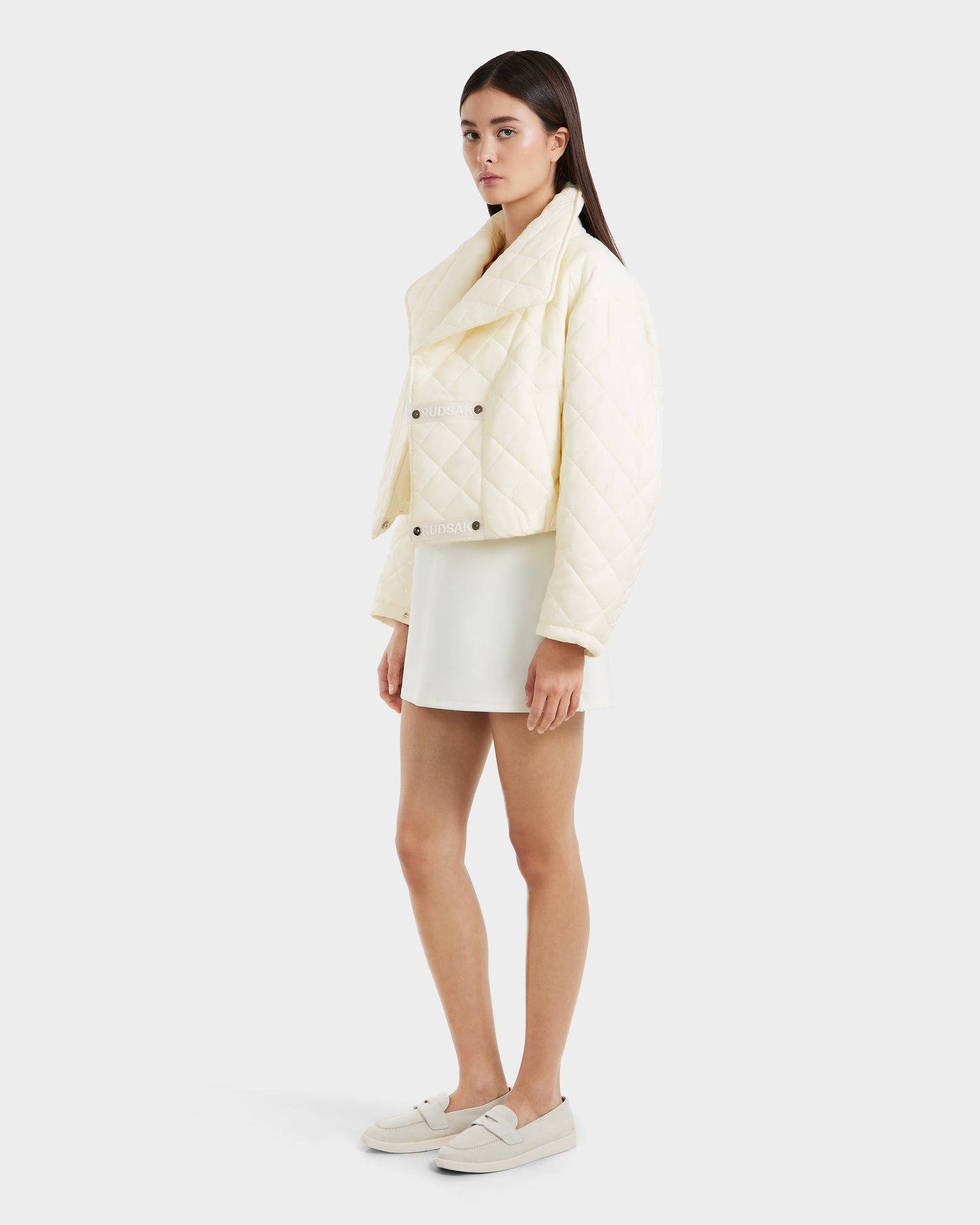 A woman with long brown hair wears the Rudsak KALEE - 8124005 Iceland cream quilted jacket, a white mini skirt, and cream loafers, posing against a plain light background.