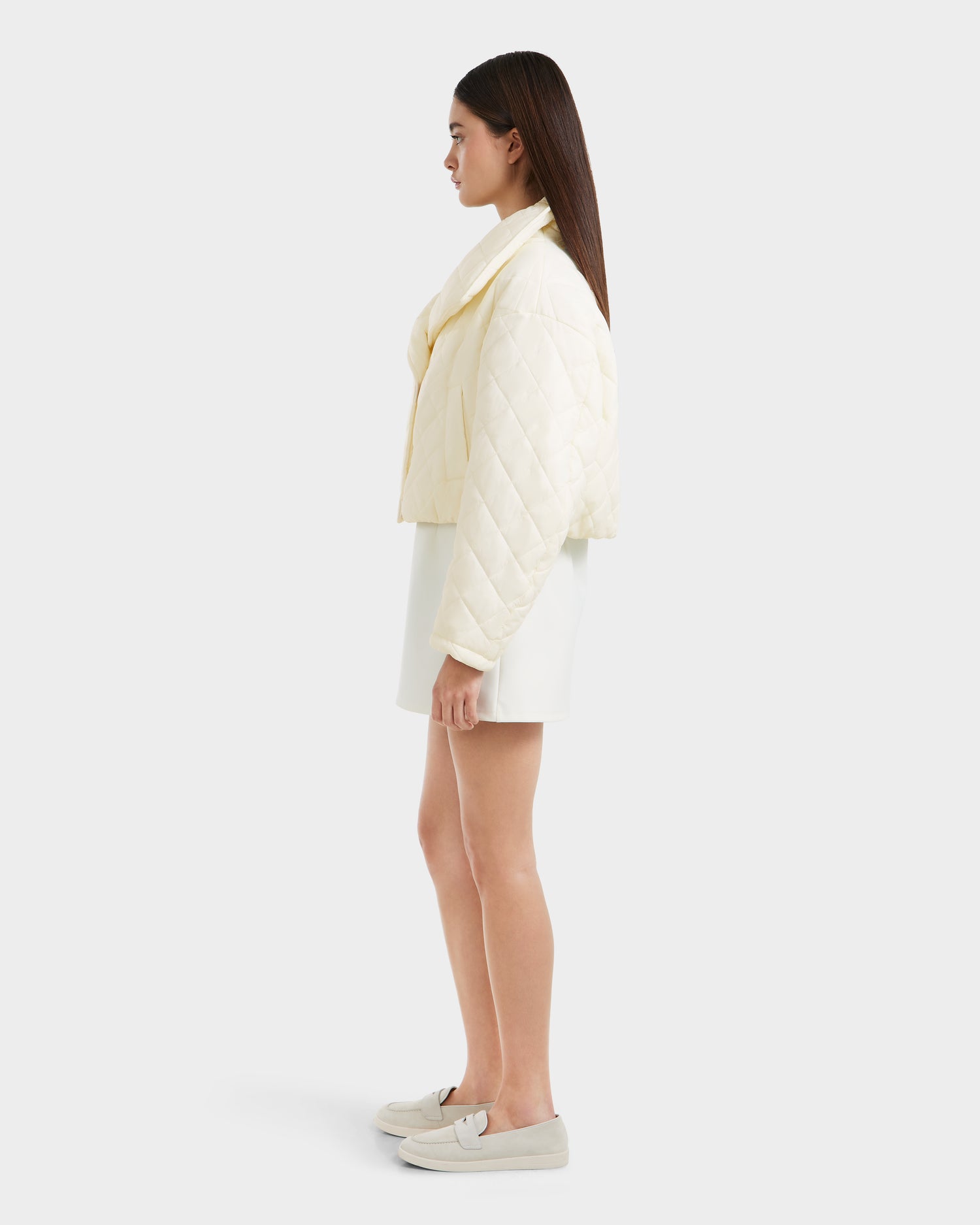 A woman stands in profile against a plain background, wearing the Rudsak KALEE - 8124005 Iceland quilted light jacket, a white skirt, and white slip-on shoes. Her straight dark hair is worn down.