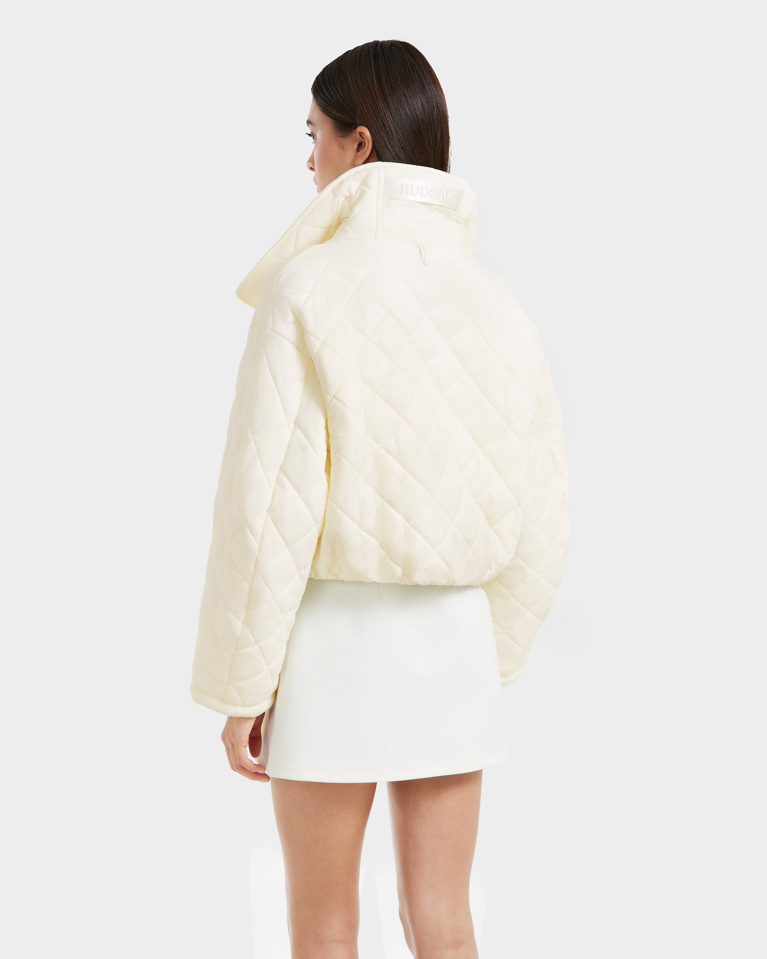 A woman with straight dark hair wears a cream KALEE - 8124005 Iceland quilted light jacket by Rudsak with a high collar and a white mini skirt, standing against a plain white background, facing away from the camera.