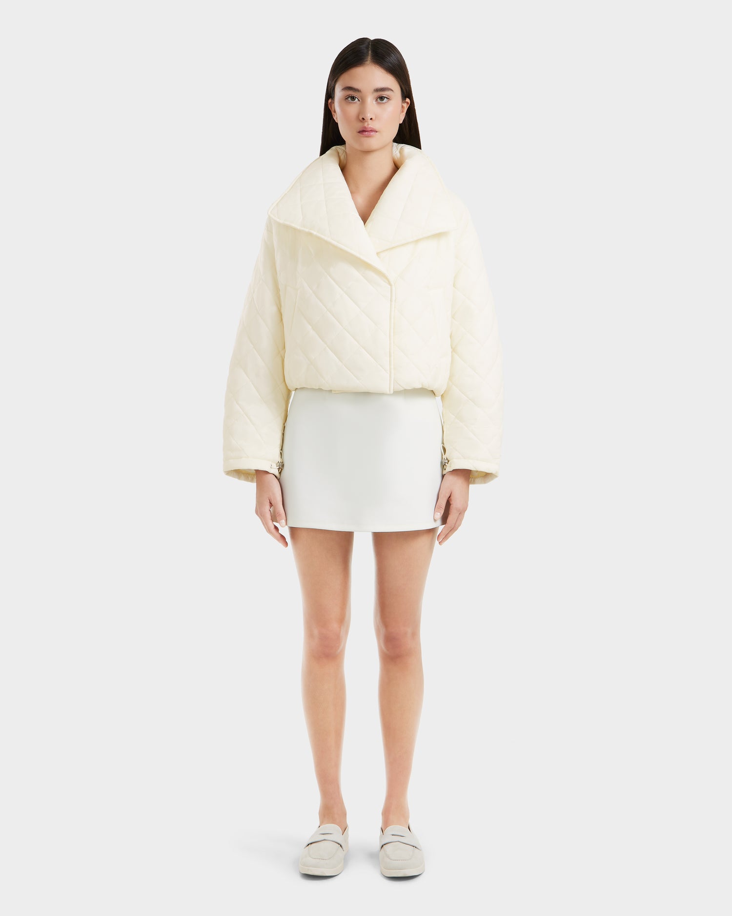 A woman faces forward wearing the Rudsak KALEE - 8124005 Iceland quilted light jacket with wide lapels, paired with a white mini skirt and slip-on shoes, set against a plain light background.