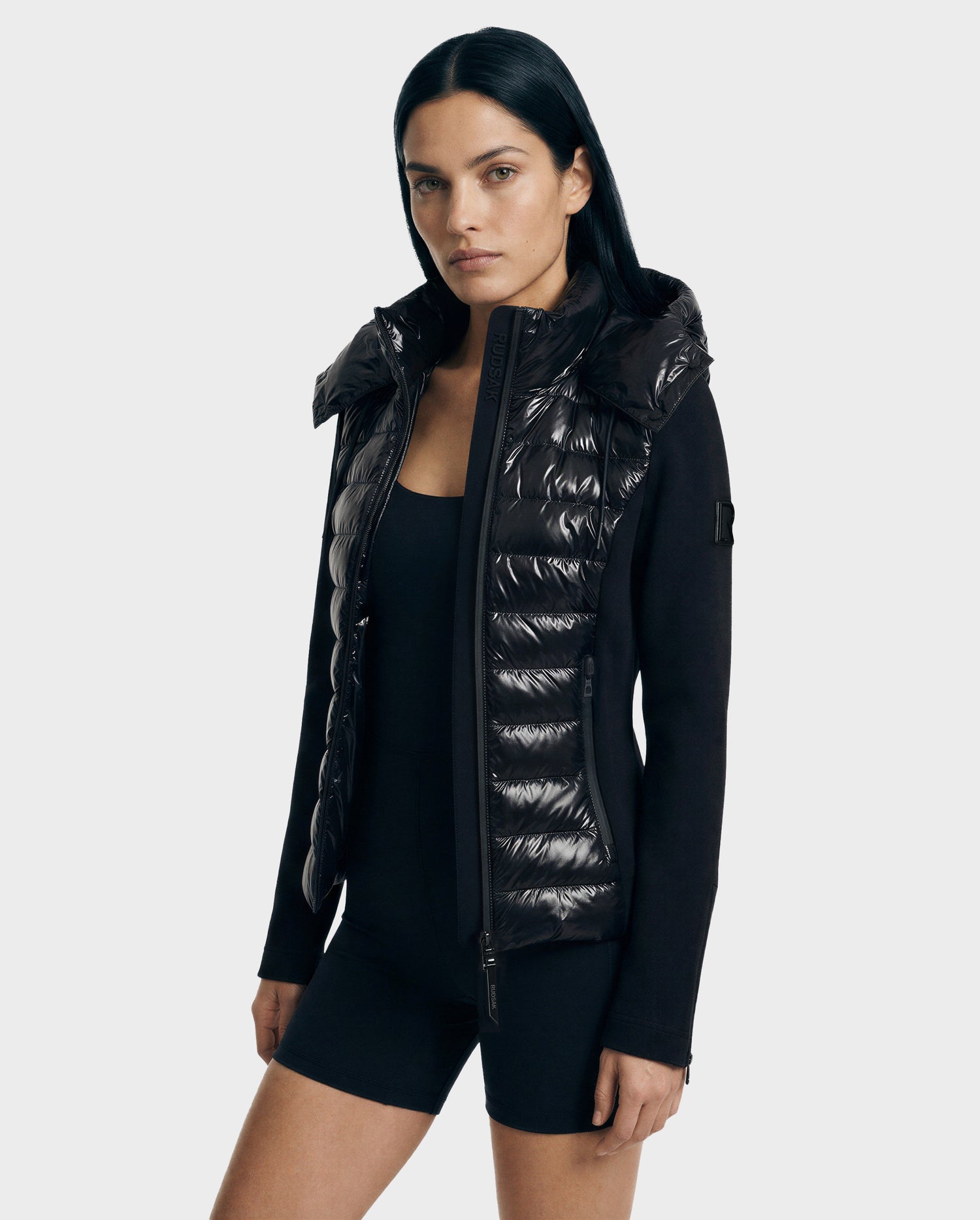 A woman with straight black hair wears the Rudsak MABS C - 8125701 Black puffer jacket with knit sleeves over a black outfit, standing against a plain light gray background.