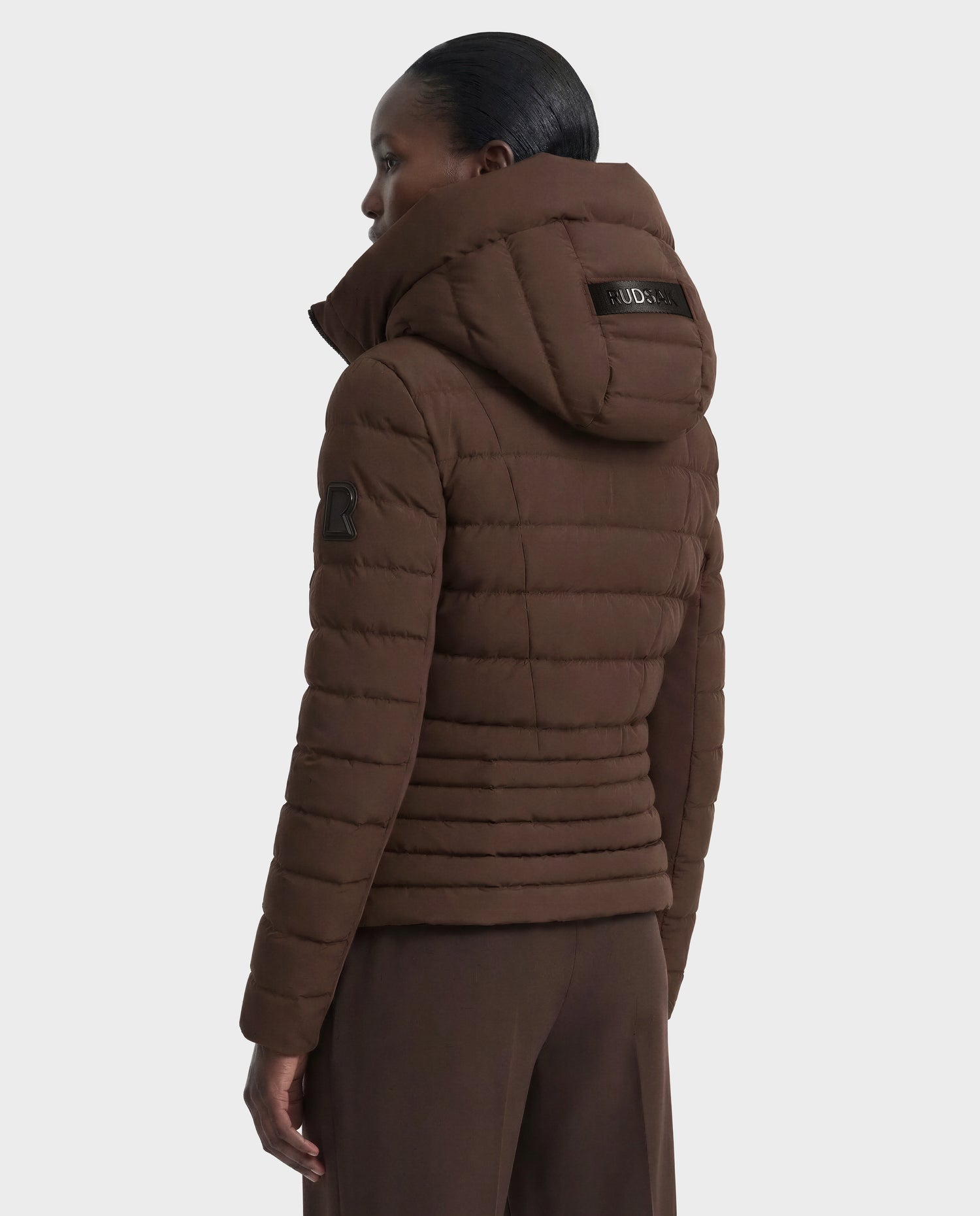A person with dark hair pulled back models the Rudsak MELISSA - 8124824 Espresso women's lightweight down puffer with hood and matching brown pants, shown from the back and side on a plain white background.
