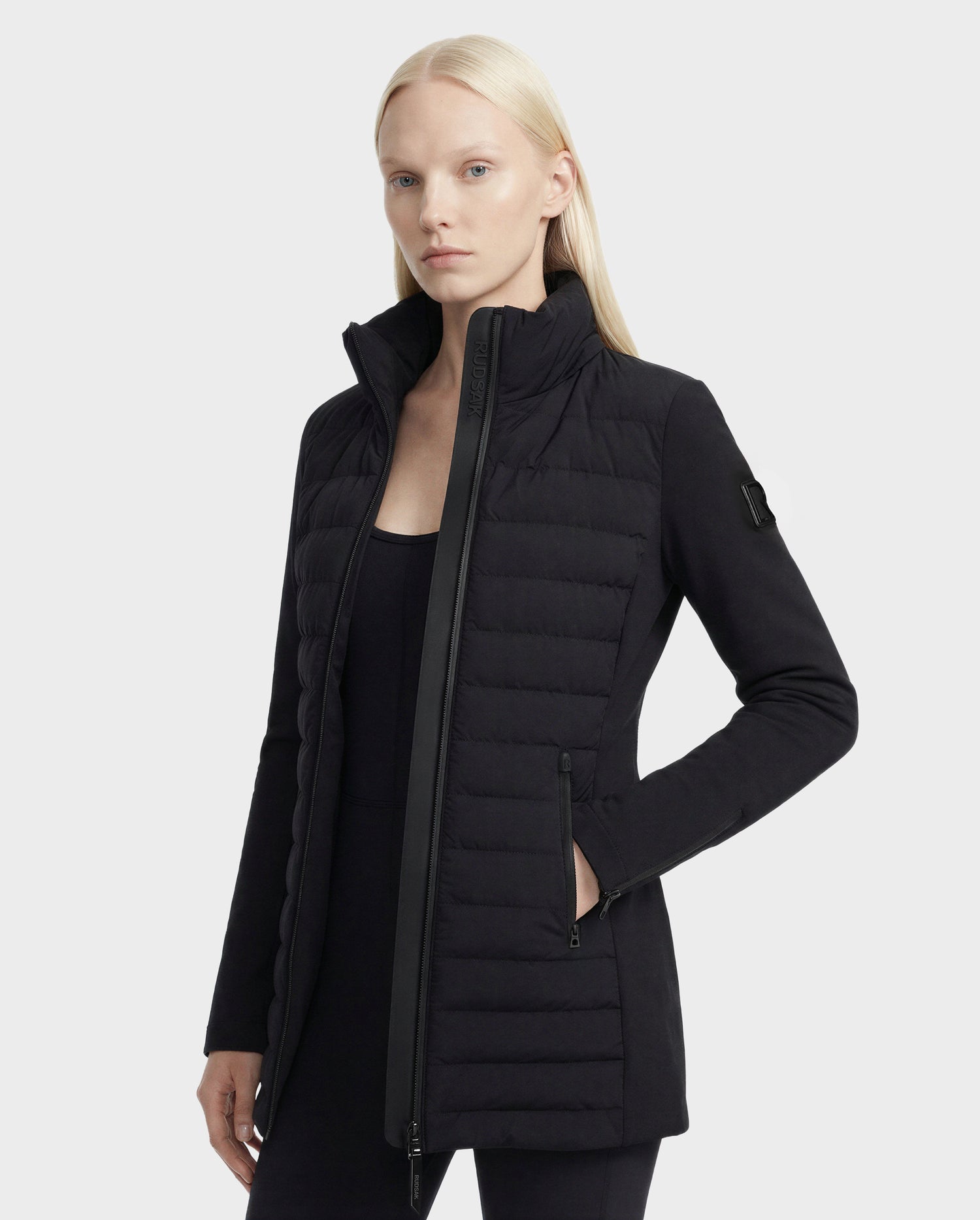 A woman with long, straight blonde hair wears the Rudsak MERINA - 8125056 Black women's quilted down jacket, featuring a high collar, zippered pockets, and a sleeve patch. She stands against a plain light grey background.