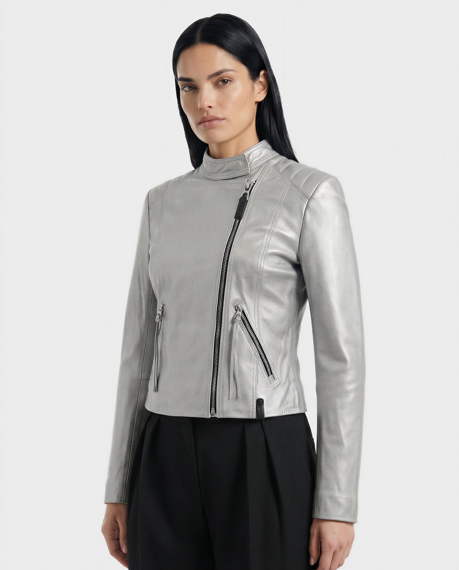 A woman with long black hair wears the Rudsak MERINA - L120013 Metallic Aluminium moto jacket, featuring an asymmetrical zipper and two front zippered pockets, paired with black pants against a plain light background.