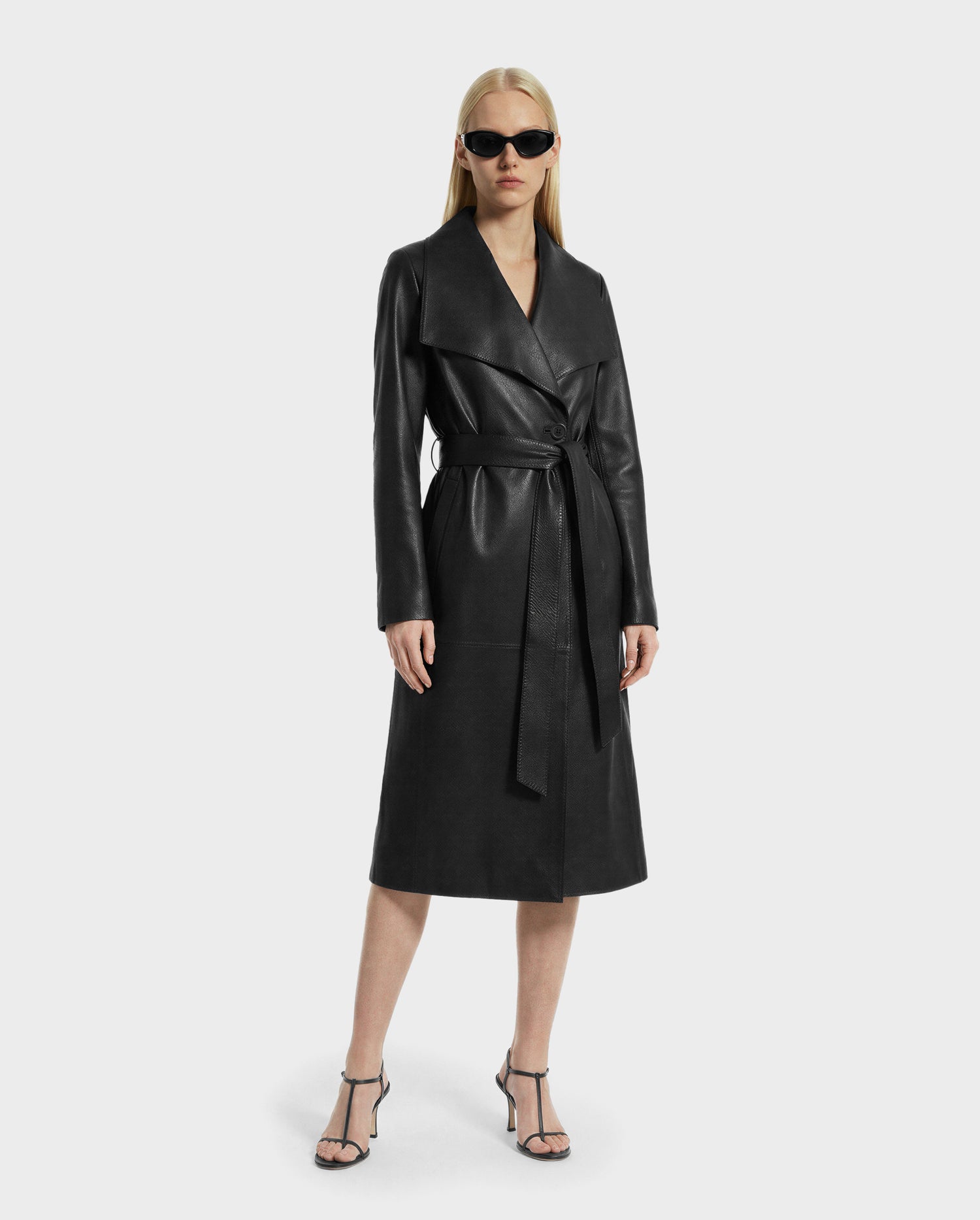 A person stands confidently against a plain light background in the MURMU X - 8125670 Black leather coat by Rudsak, styled with black sunglasses and strappy black heels—a striking example of investment dressing.