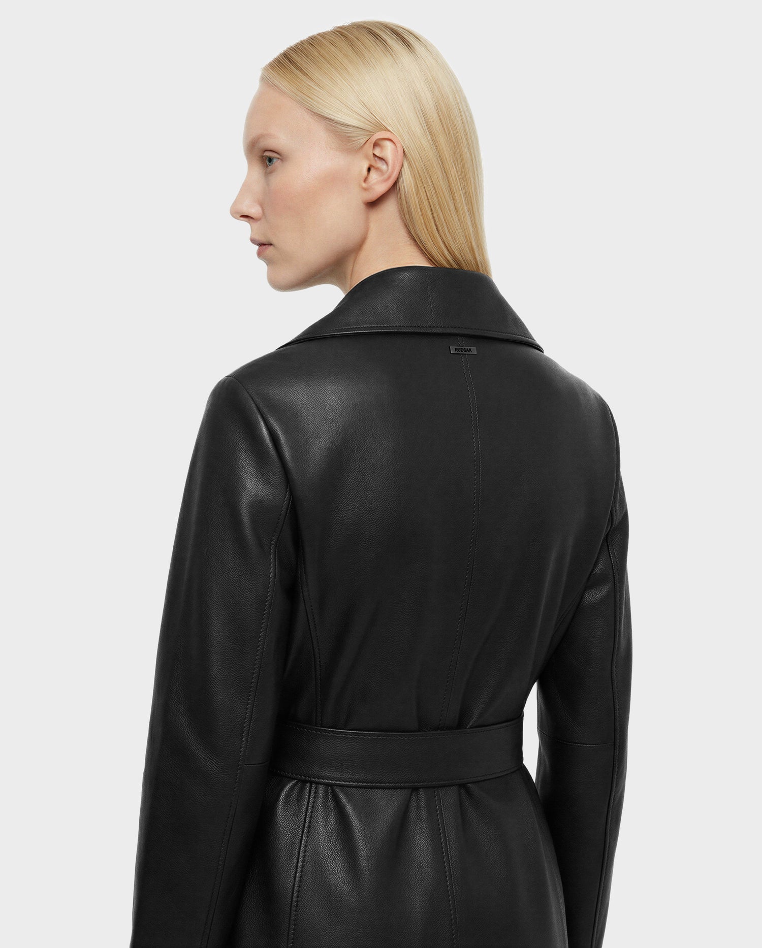 A woman with straight blonde hair, seen from the back and side, wears the Rudsak MURMU X - 8125670 Black top-grain leather belted coat with a wide collar against a plain light gray background.