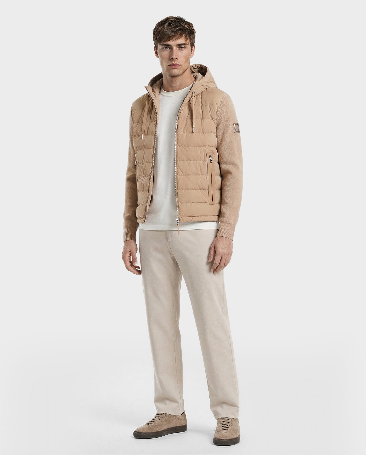 A man faces forward wearing the Rudsak RIVER - 6124584 Almond men’s lightweight down jacket with a hood, paired with a white shirt, beige trousers, and beige sneakers against a plain white background.