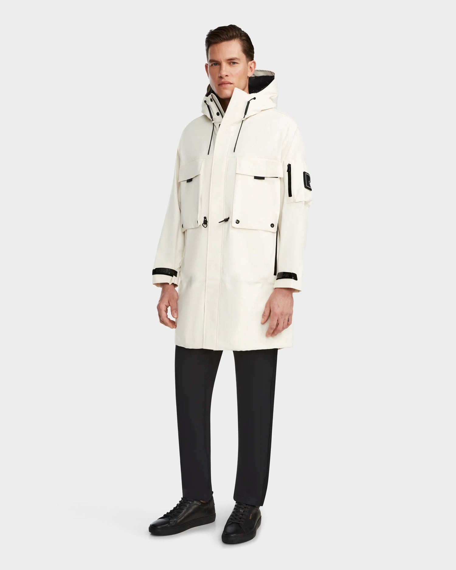 A man wearing the RUDSAK MELI - 9123003 ICELAND long white unisex raincoat with multiple pockets, black trousers, and black sneakers stands against a plain light gray background.