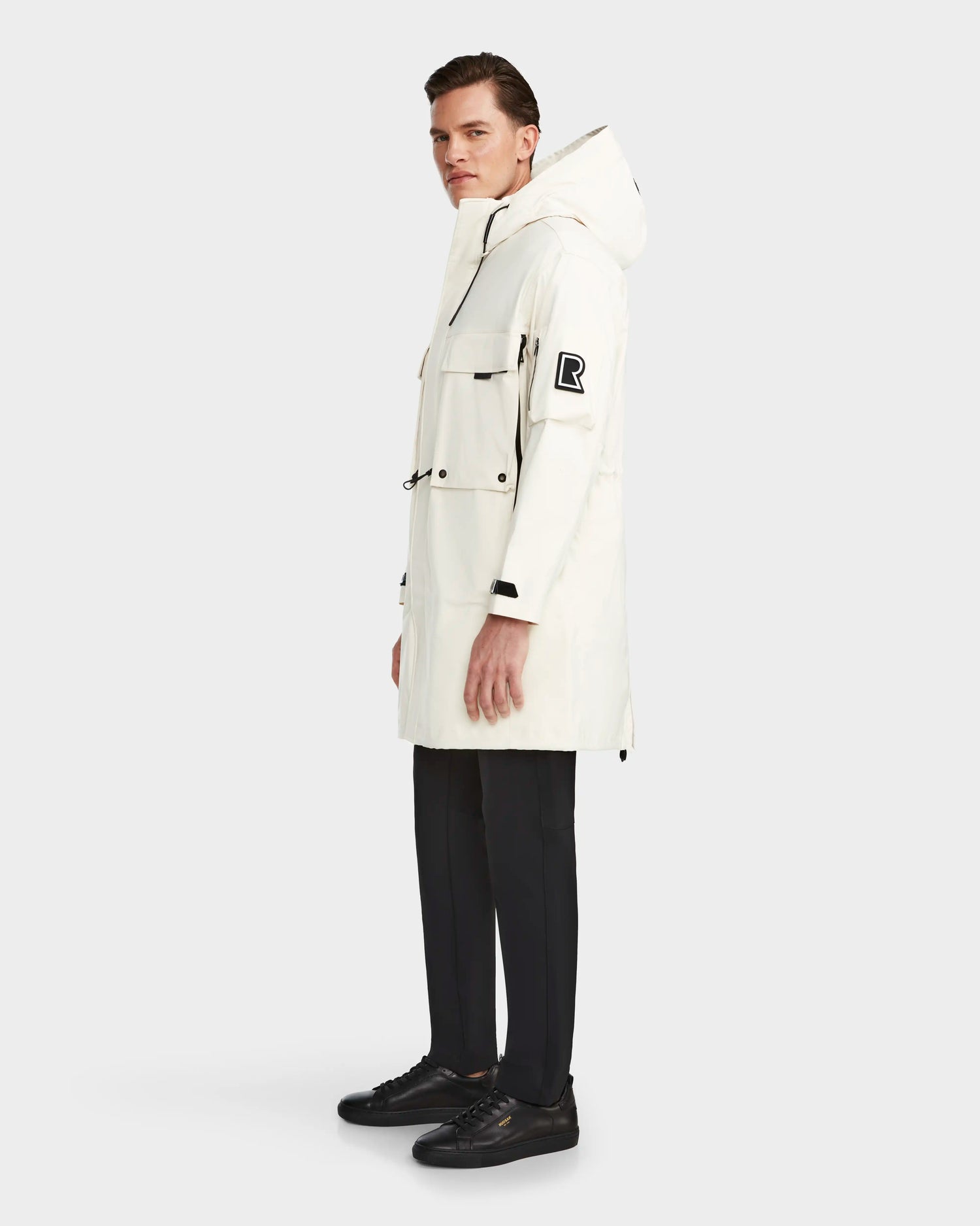 A man stands slightly to the side against a plain white background wearing the RUDSAK MELI - 9123003 ICELAND long white unisex raincoat, featuring black details, a large R patch on the upper sleeve, and made from recycled Stormshell fabric.