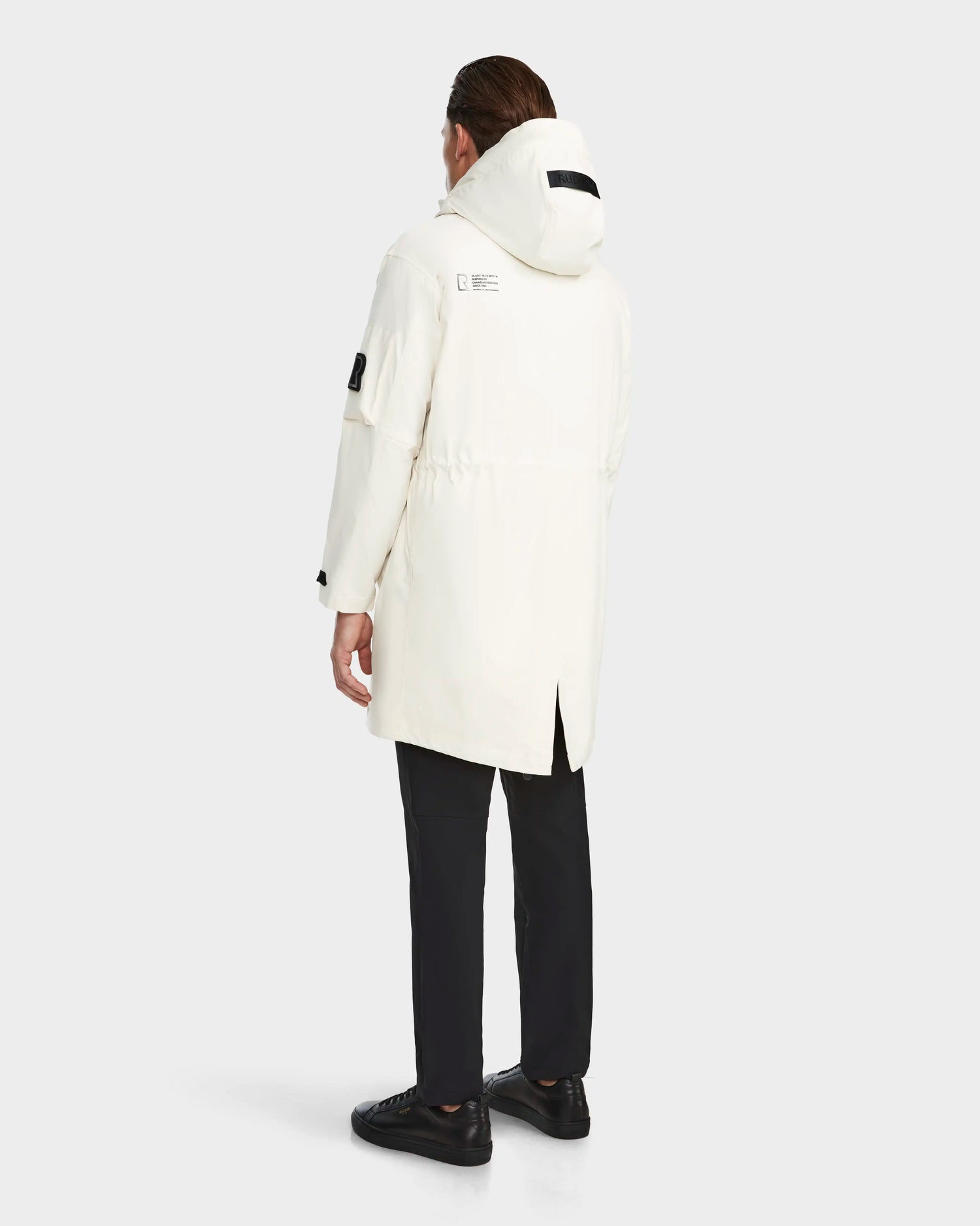A person faces away, wearing the RUDSAK MELI - 9123003 ICELAND long unisex white raincoat made from recycled Stormshell fabric, paired with black pants and shoes, set against a plain light gray background.