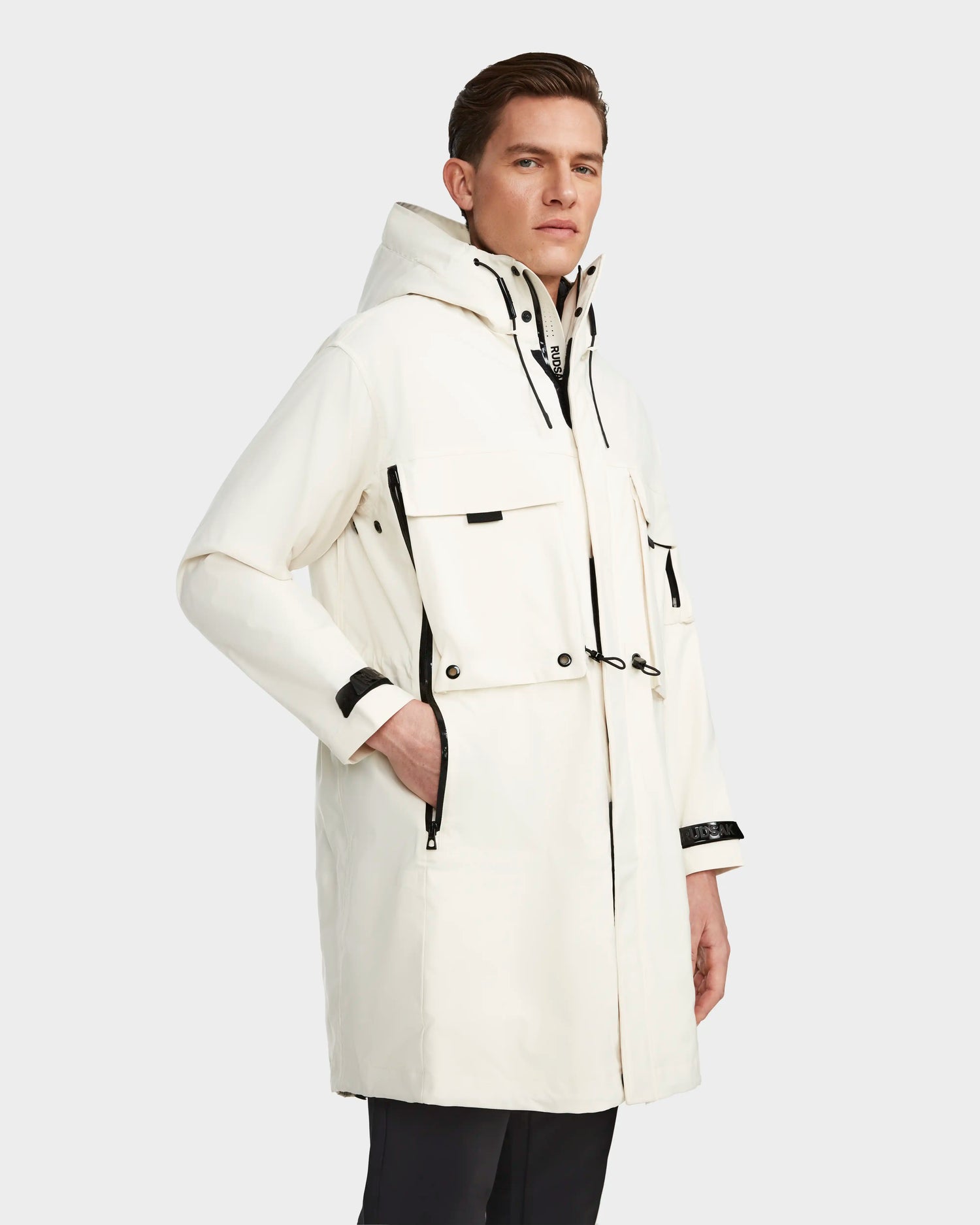 A man stands with one hand in his pocket against a light gray background, wearing the RUDSAK MELI - 9123003 ICELAND long white unisex raincoat crafted from recycled Stormshell fabric with black zipper and button accents.