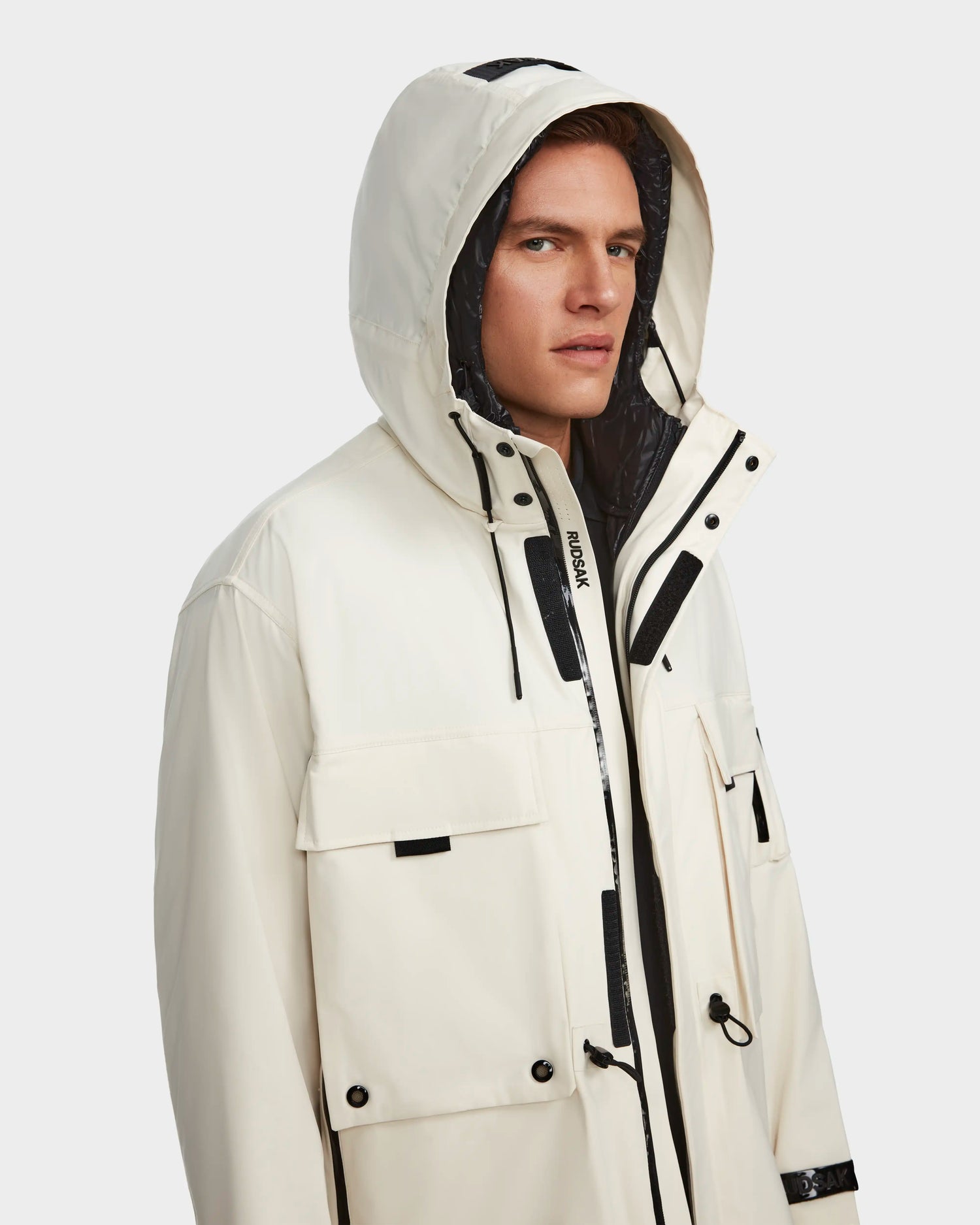 A man wears the RUDSAK MELI - 9123003 ICELAND unisex raincoat in white with black accents, front pockets, and a high collar, made from recycled Stormshell fabric, standing against a plain background and looking slightly to the side.