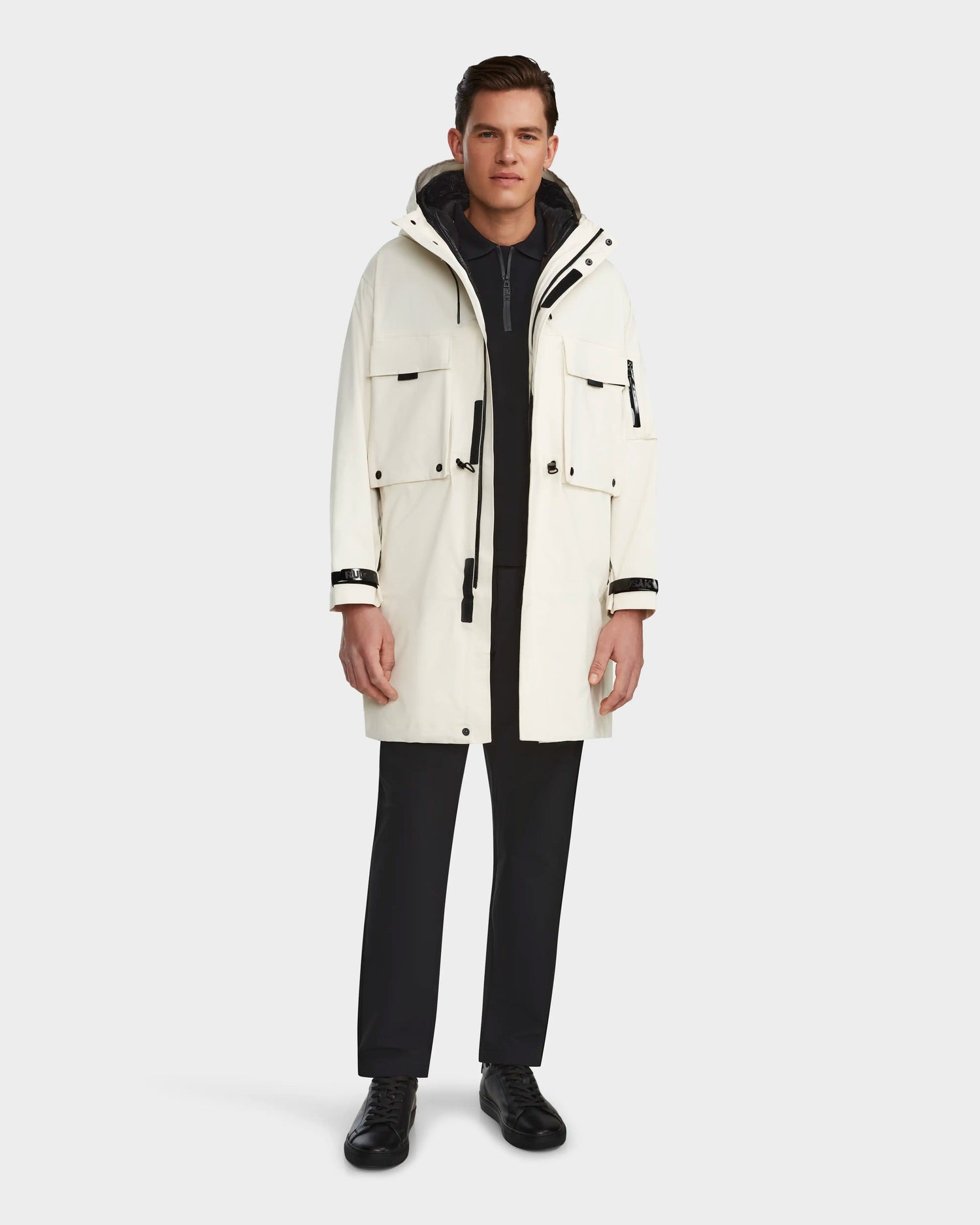 A person stands facing forward against a plain light background, wearing the RUDSAK MELI - 9123003 ICELAND long white unisex raincoat with black details, zipped over a dark outfit.
