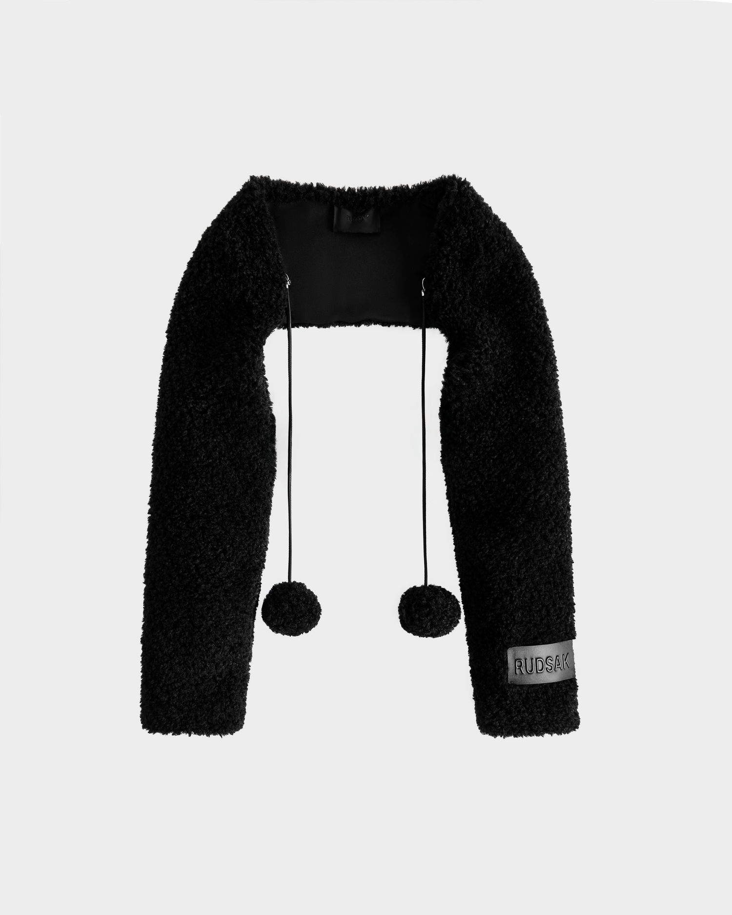 The VERITY - 8525666 Black by Rudsak is a black, textured cropped shrug with long sleeves, two pom-pom drawstrings, and a RUBACK patch on the left wrist—ideal as a chic winter accessory.
