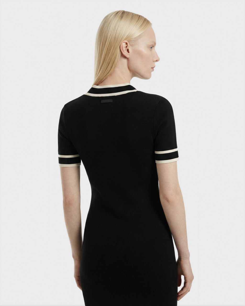 A woman with straight blonde hair is seen from behind wearing the Rudsak VINNIE - 8526038 Black fitted knit midi dress, featuring white trim on the sleeves and collar, against a plain light background.
