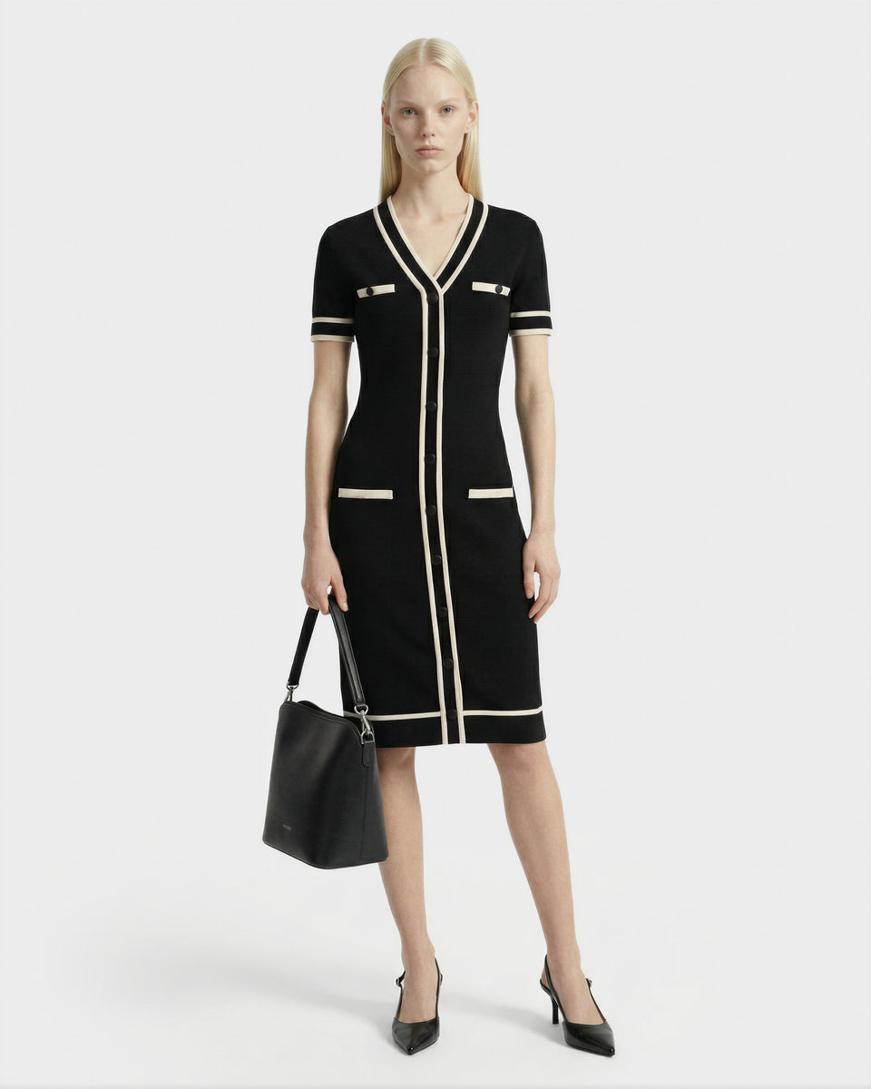 A woman with long blonde hair models the VINNIE - 8526038 Black knit midi dress by Rudsak with white trim, styled with black pointed-toe heels and a black handbag, posing against a plain white background.
