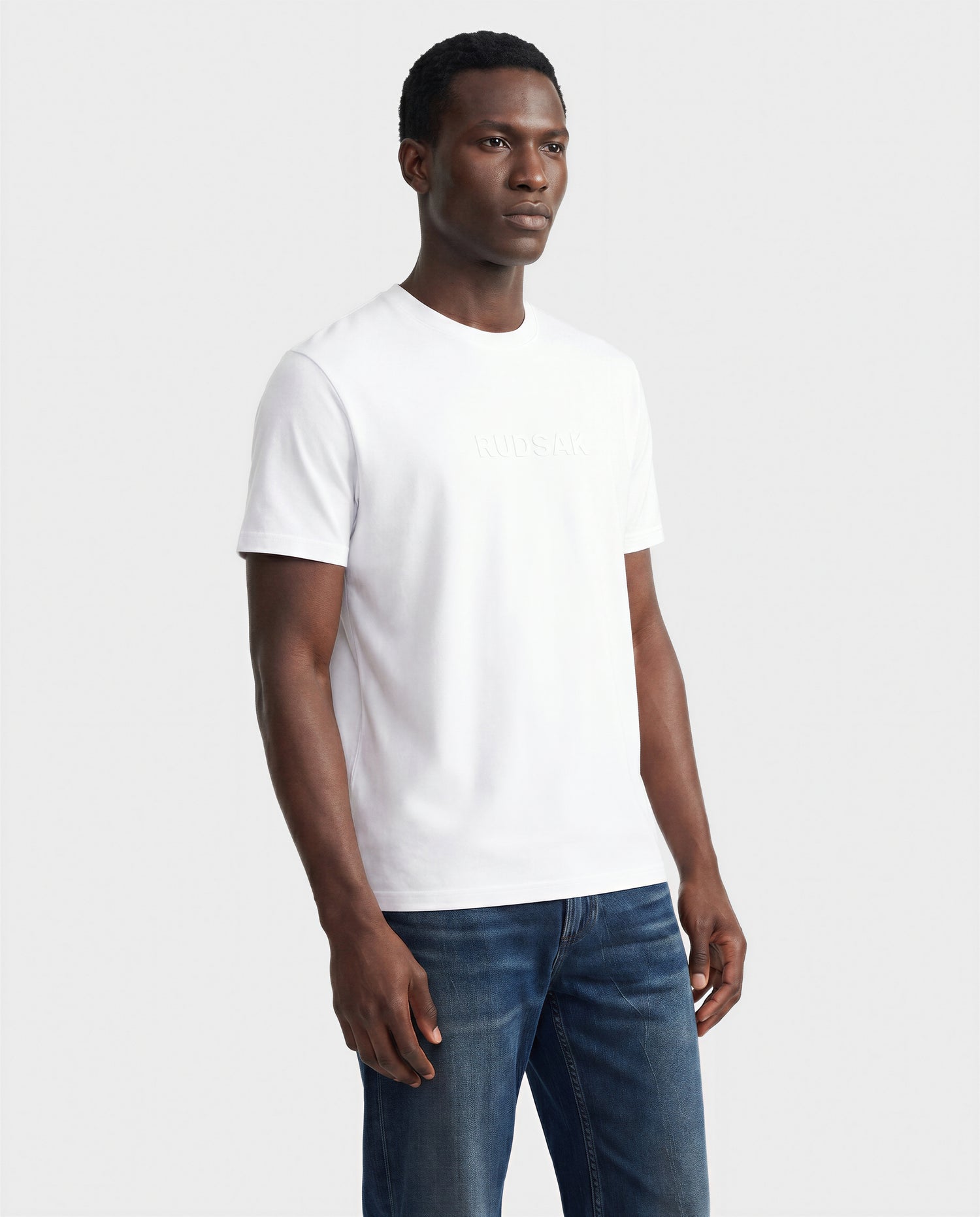 A man stands against a plain white background, wearing the Rudsak FERDI - 6522005 Natural White short-sleeve t-shirt with subtle embossed text and blue jeans, facing slightly left with a neutral expression.