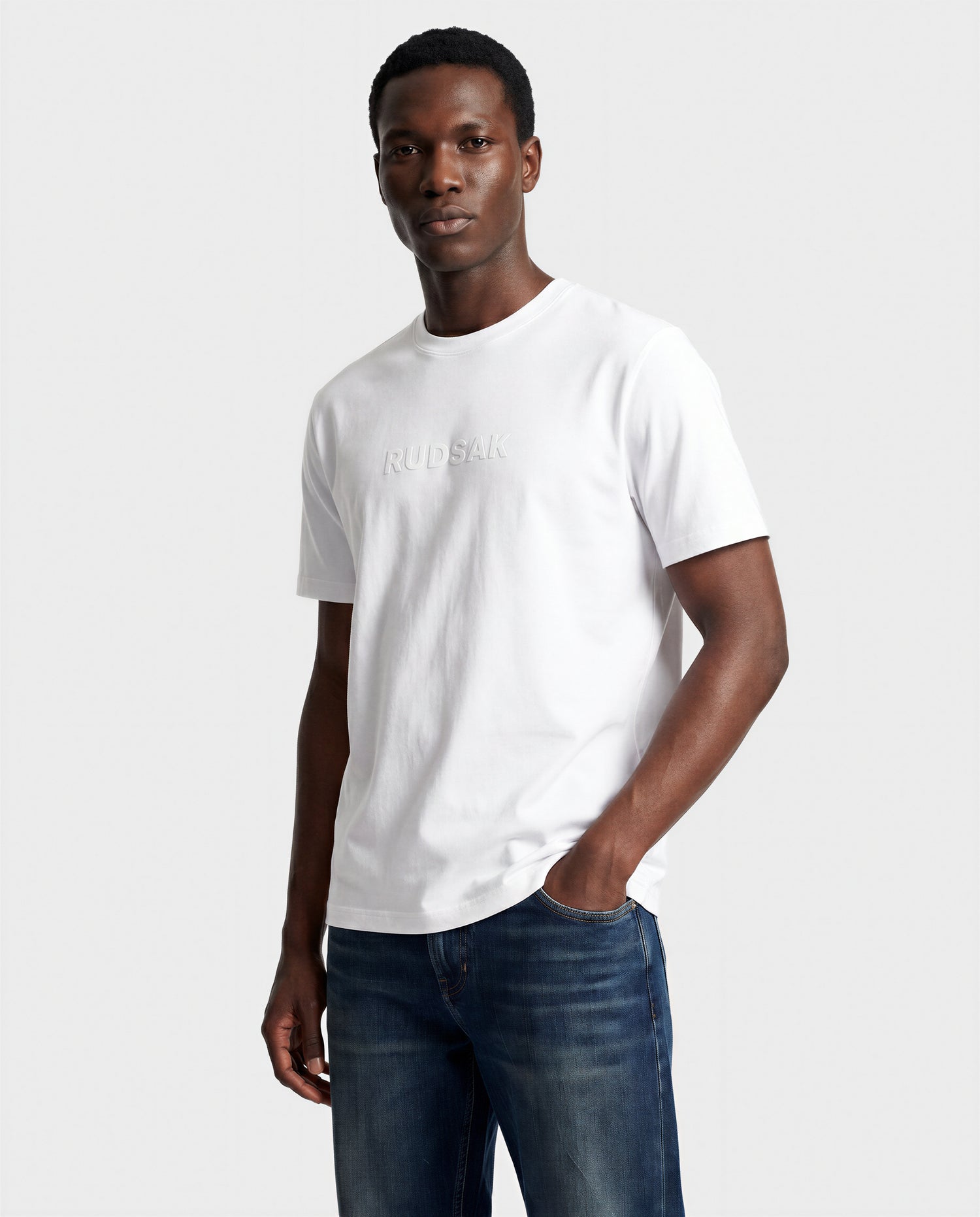 A man in the Rudsak FERDI - 6522005 Natural White T-shirt and blue jeans stands against a light gray background, looking at the camera with one hand in his pocket.