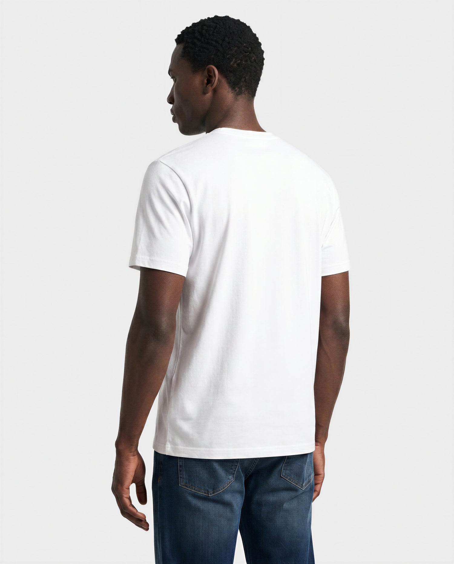 A man stands facing away, wearing the Rudsak FERDI - 6522005 Natural White short-sleeve t-shirt and blue jeans against a light gray background.