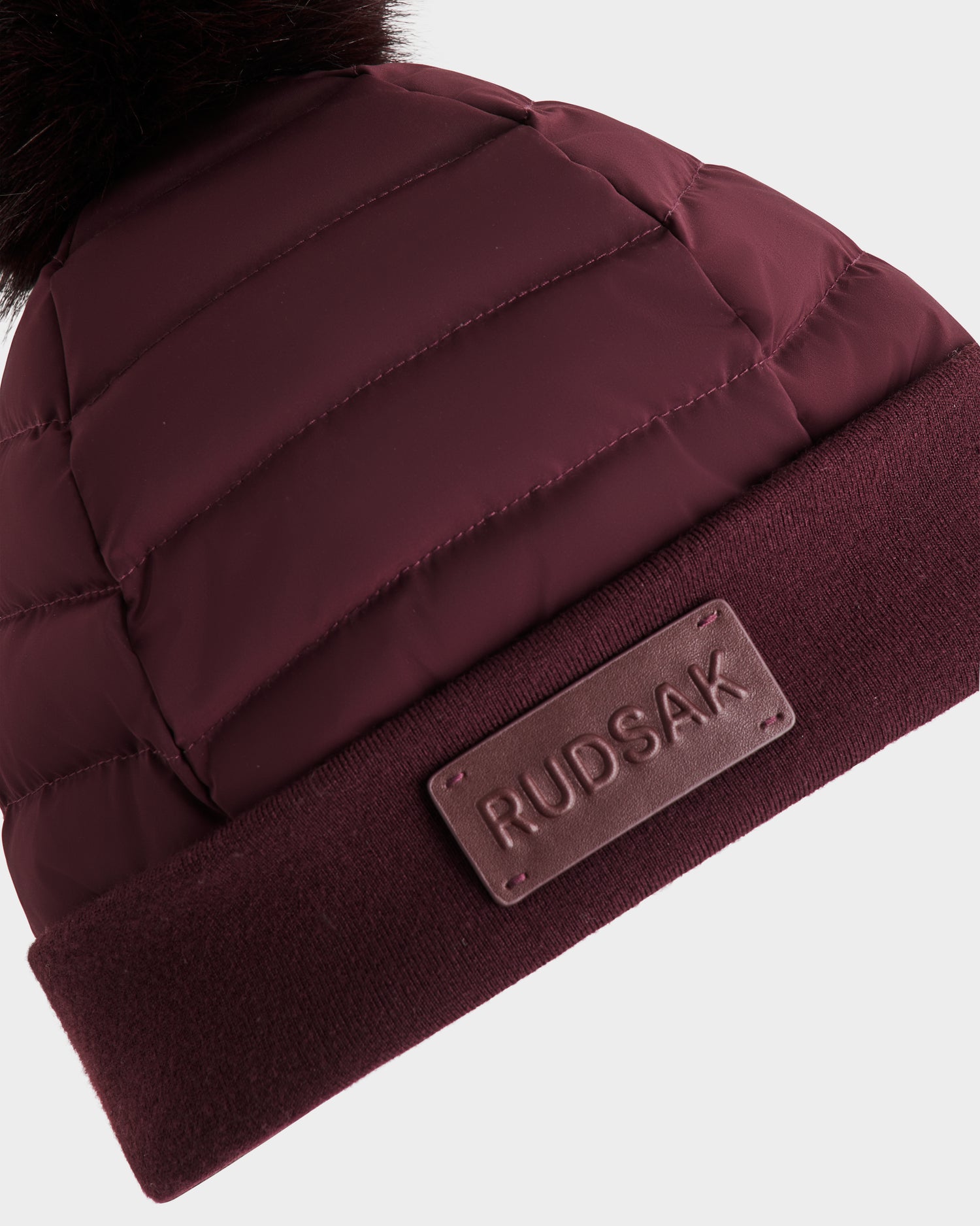 Close-up of the GALA - 8424514 Bordeaux womens down puffer hat by Rudsak, featuring a padded quilted texture, knit fold-over cuff, leather Rudsak patch, and a removable faux fur pom pom for versatile style.