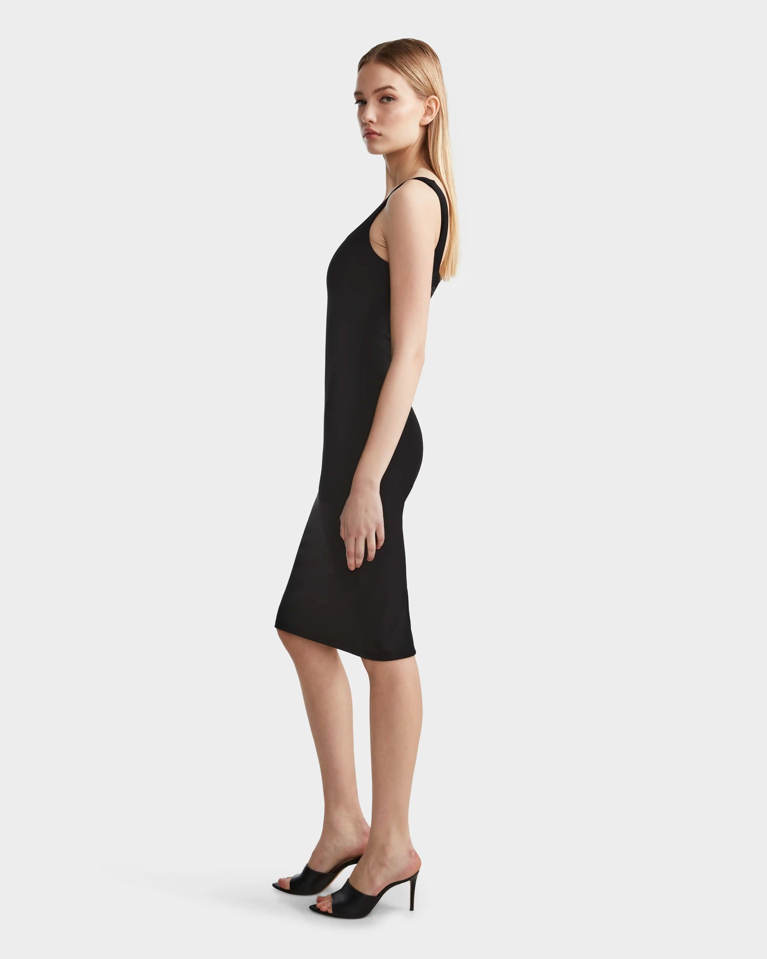 A woman with straight blonde hair wears the Rudsak SIMONE - 8524065 Black fitted sleeveless midi dress and black heeled mules, standing sideways against a plain white background.