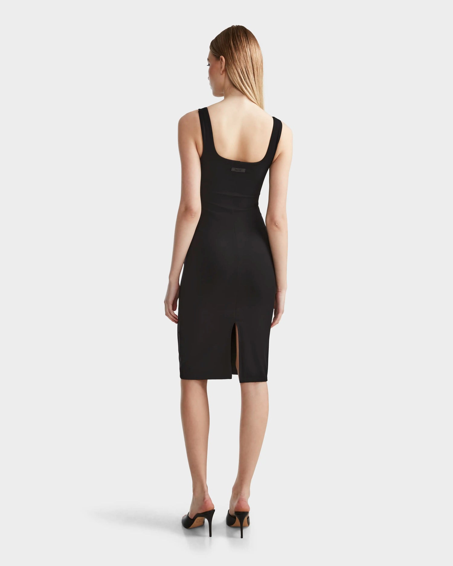 A woman with straight, light brown hair models the Rudsak SIMONE - 8524065 Black dress, a fitted sleeveless knee-length piece with a back slit, paired with black heeled sandals. She stands with her back to the camera against a plain background.