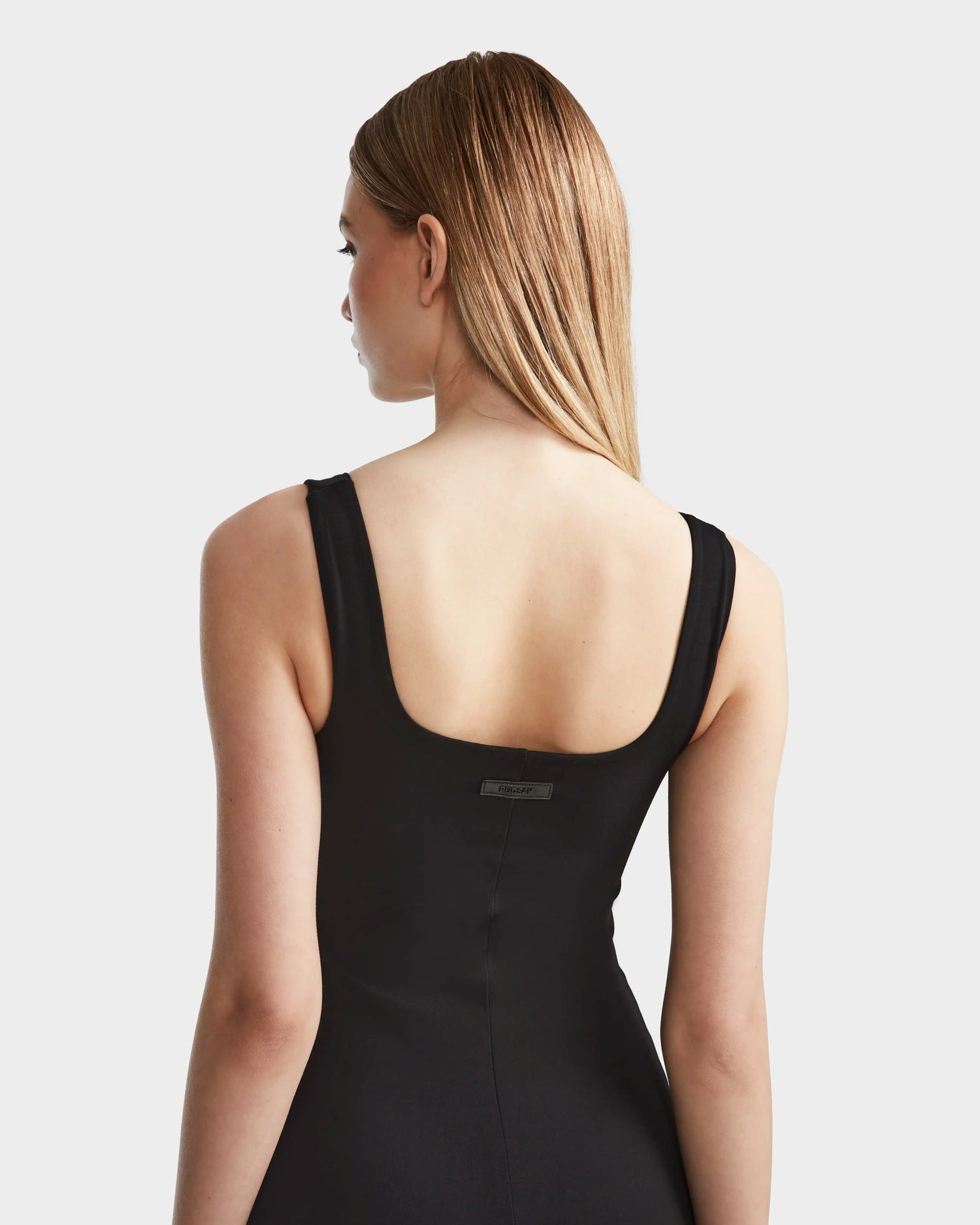 A woman with straight, light brown hair is shown from the back wearing the SIMONE - 8524065 Black sleeveless midi dress by Rudsak, featuring a square neckline and low back. The background is plain light gray.