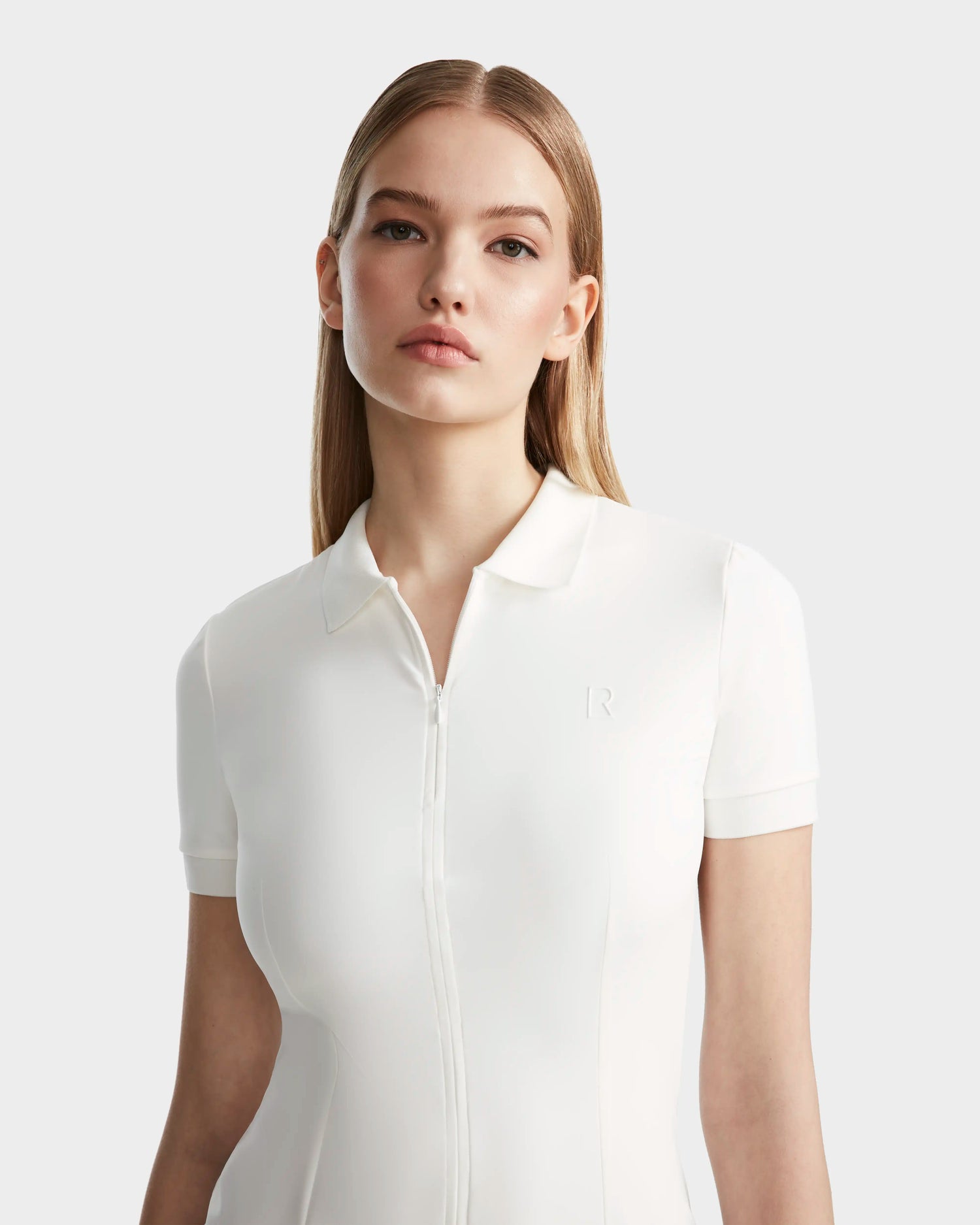 A young woman with straight, light brown hair models the Rudsak SUKI - 8524029 Cotton White women's polo dress, featuring short sleeves, a front zipper, and a subtle R logo, standing against a plain light grey background.