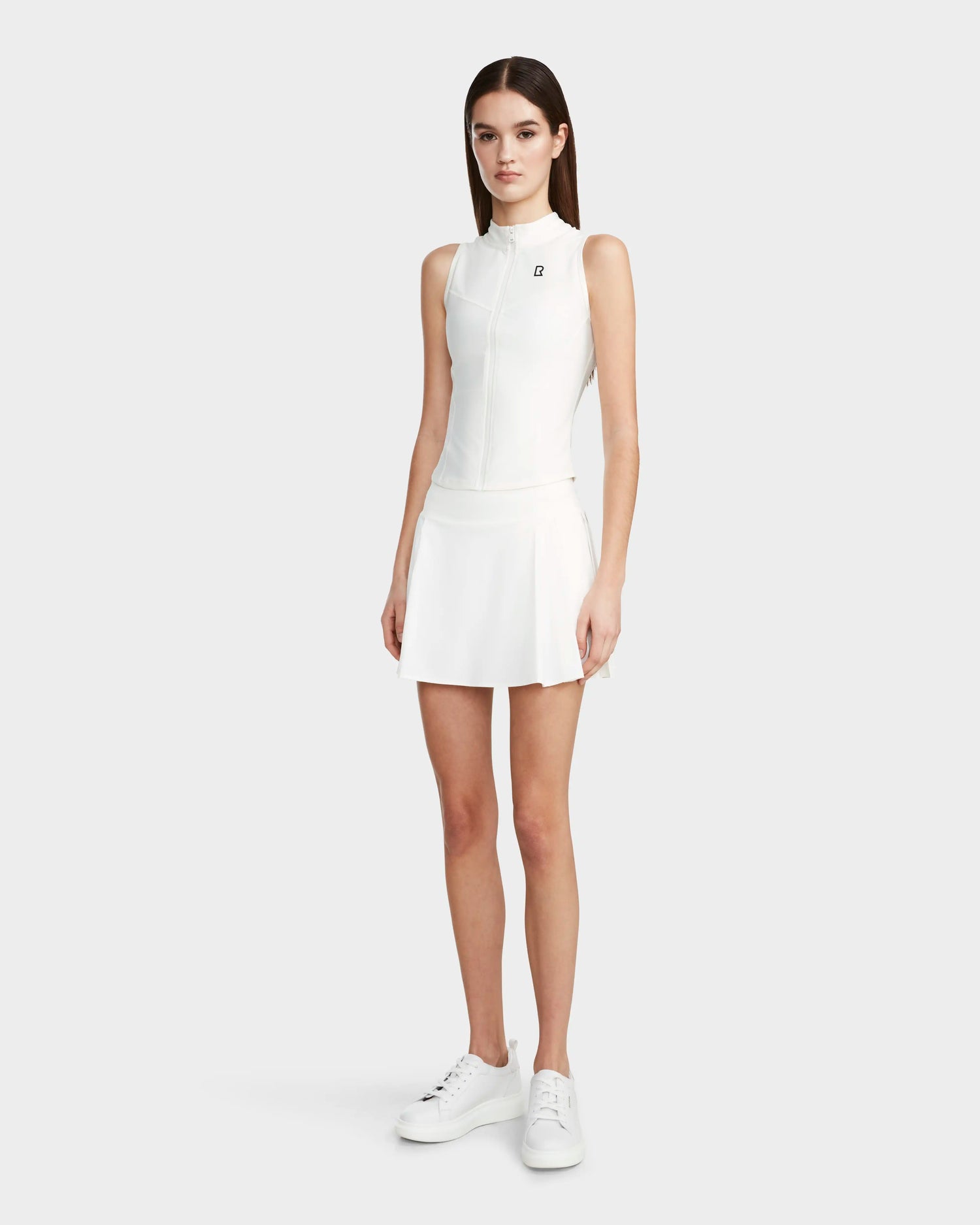 A woman with straight brown hair and a neutral expression wears the Rudsak PATTY - 8524073 Eggshell fitted zip-up sleeveless top, paired with a white pleated skirt and white sneakers, standing against a plain light background.