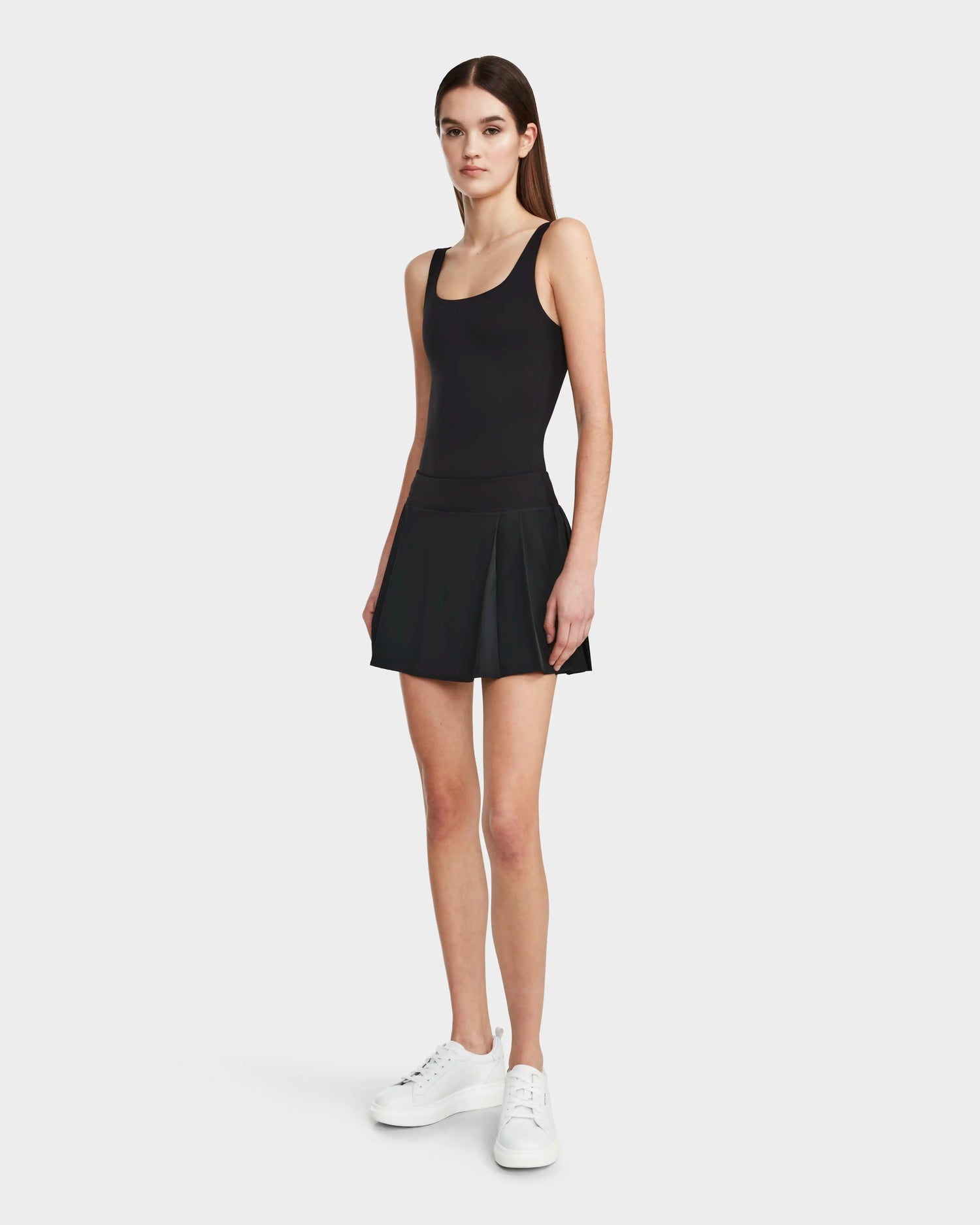 A young woman stands facing forward in a sleeveless HILDA - 8524034 Black top by Rudsak, paired with a black pleated tennis skirt and white sneakers, set against a plain light background.