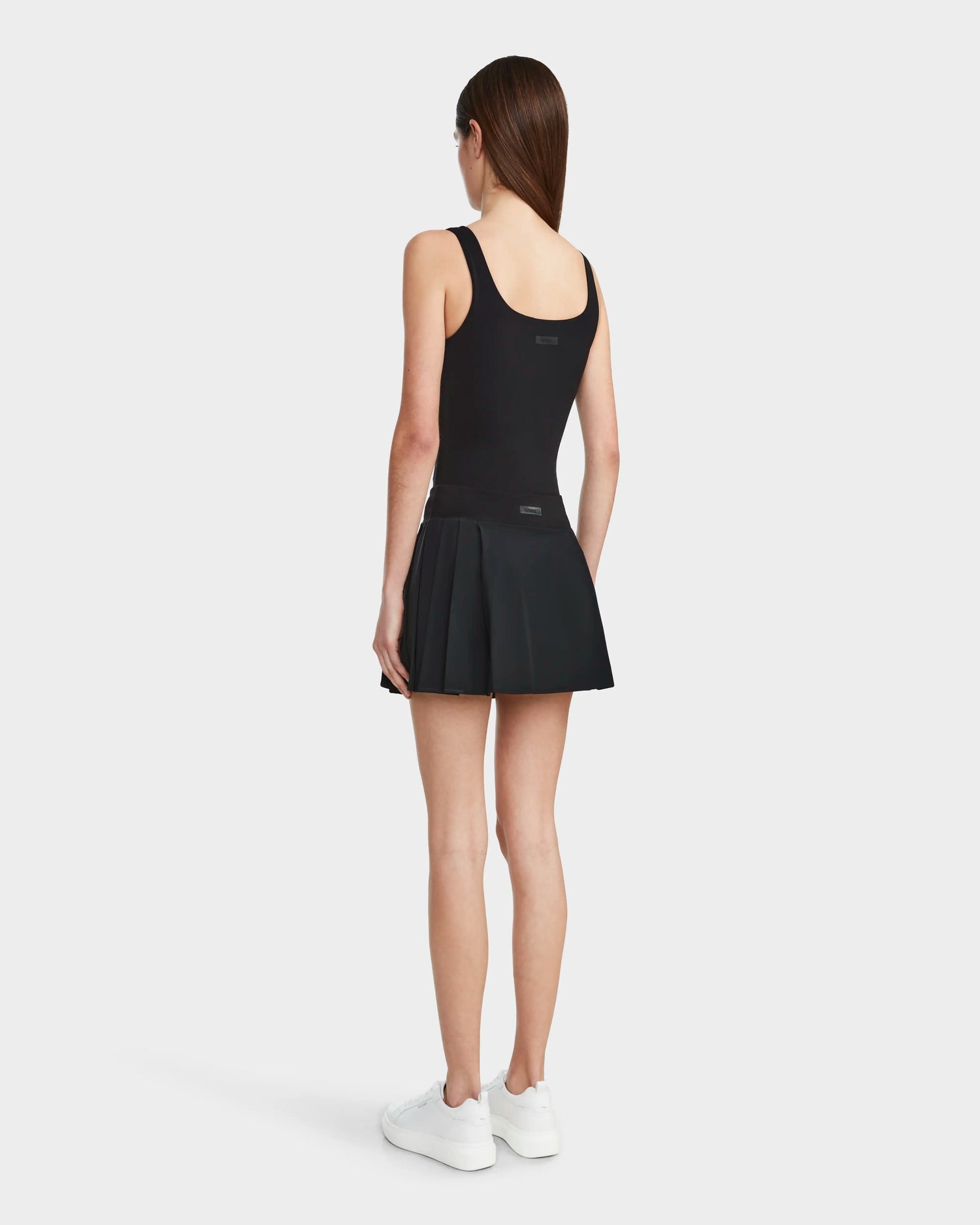 A woman stands facing away, wearing the Rudsak HILDA - 8524034 Black sleeveless top with a sporty pleated tennis skirt and white sneakers, set against a plain white background.