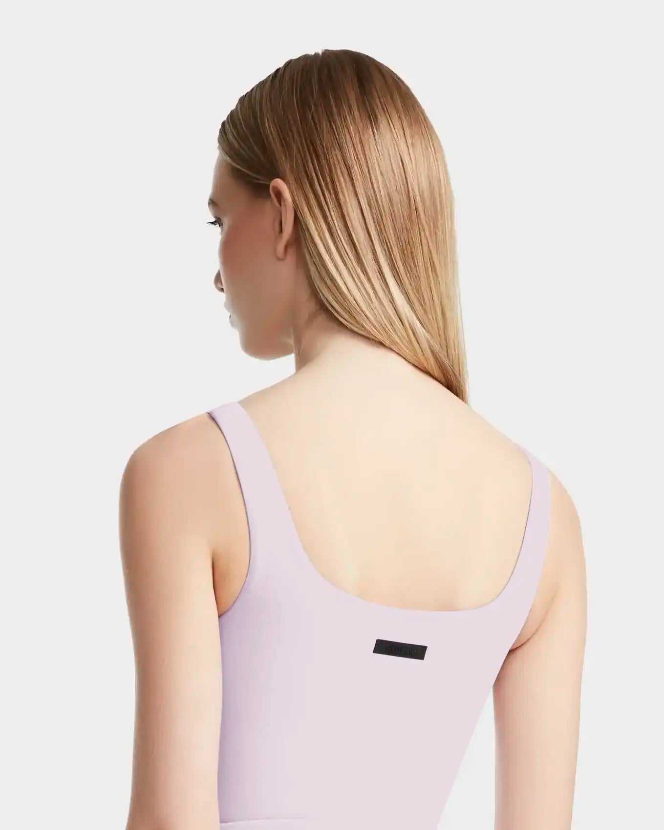 A woman with straight, light brown hair is seen from the back wearing the Rudsak BRIDGET - 8524033 Lilac Foam sleeveless tank bodysuit, which features a black rectangular tag near the neckline. The background is plain and light gray.