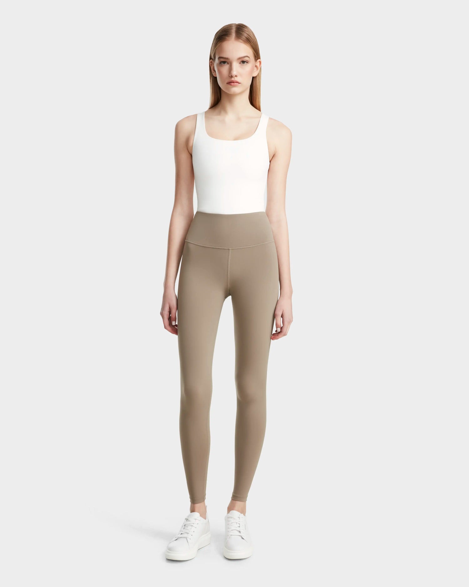 A woman faces forward in a white tank top, Rudsak’s RIANA - 8524031 Cement taupe stretch leggings, and white sneakers against a light gray background—the ideal athleisure look.